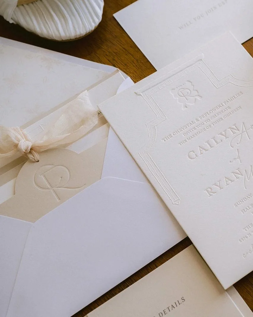 Wedding invitations and envelopes on a wooden surface, with embossed and printed text, some with decorative borders and a tied envelope with a monogram letter 'P'.