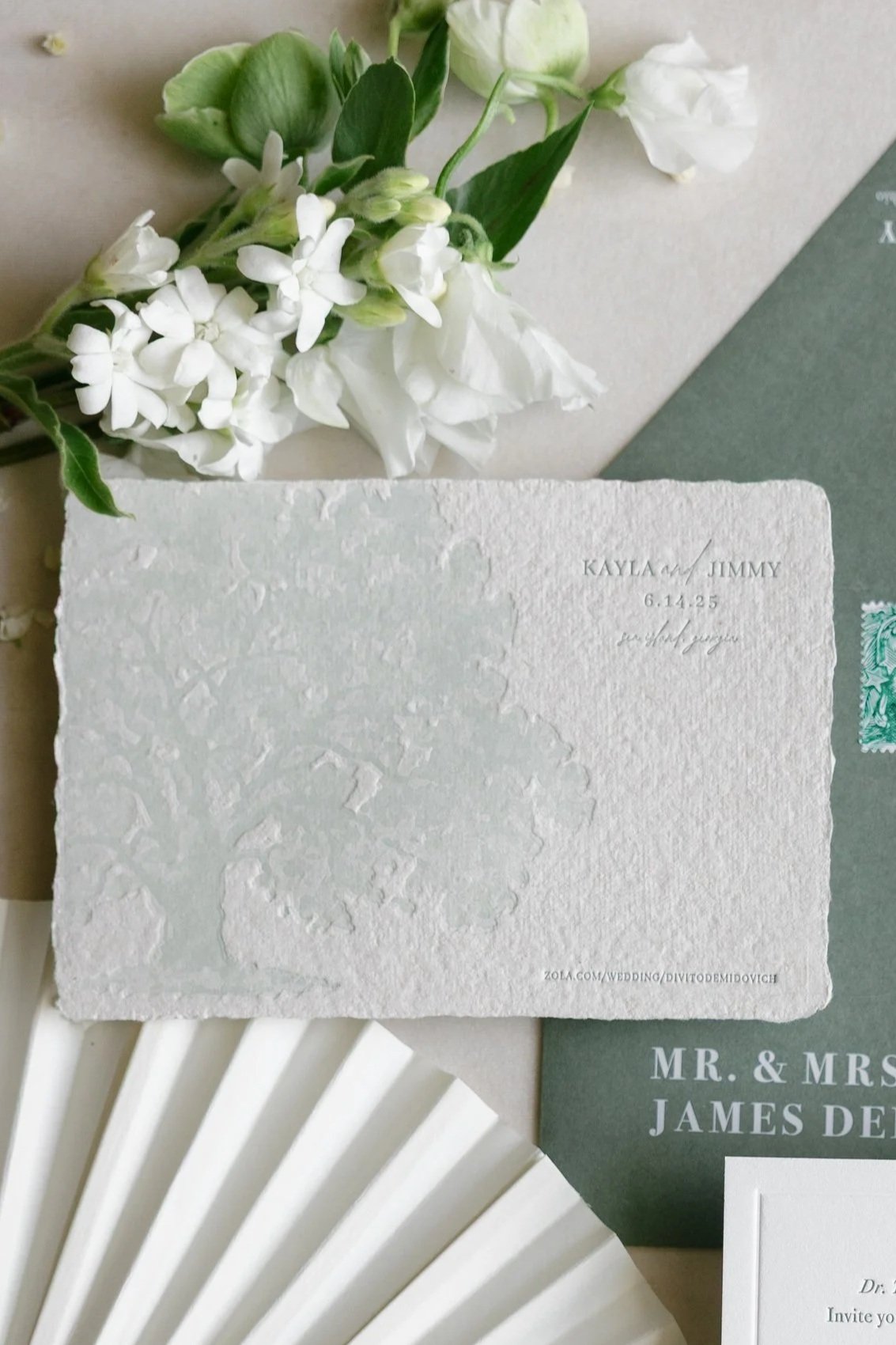 Close-up of a textured white wedding invitation card with embossed details, partially visible greenery and white flowers in the background, and a green antique stamp on a green backing.