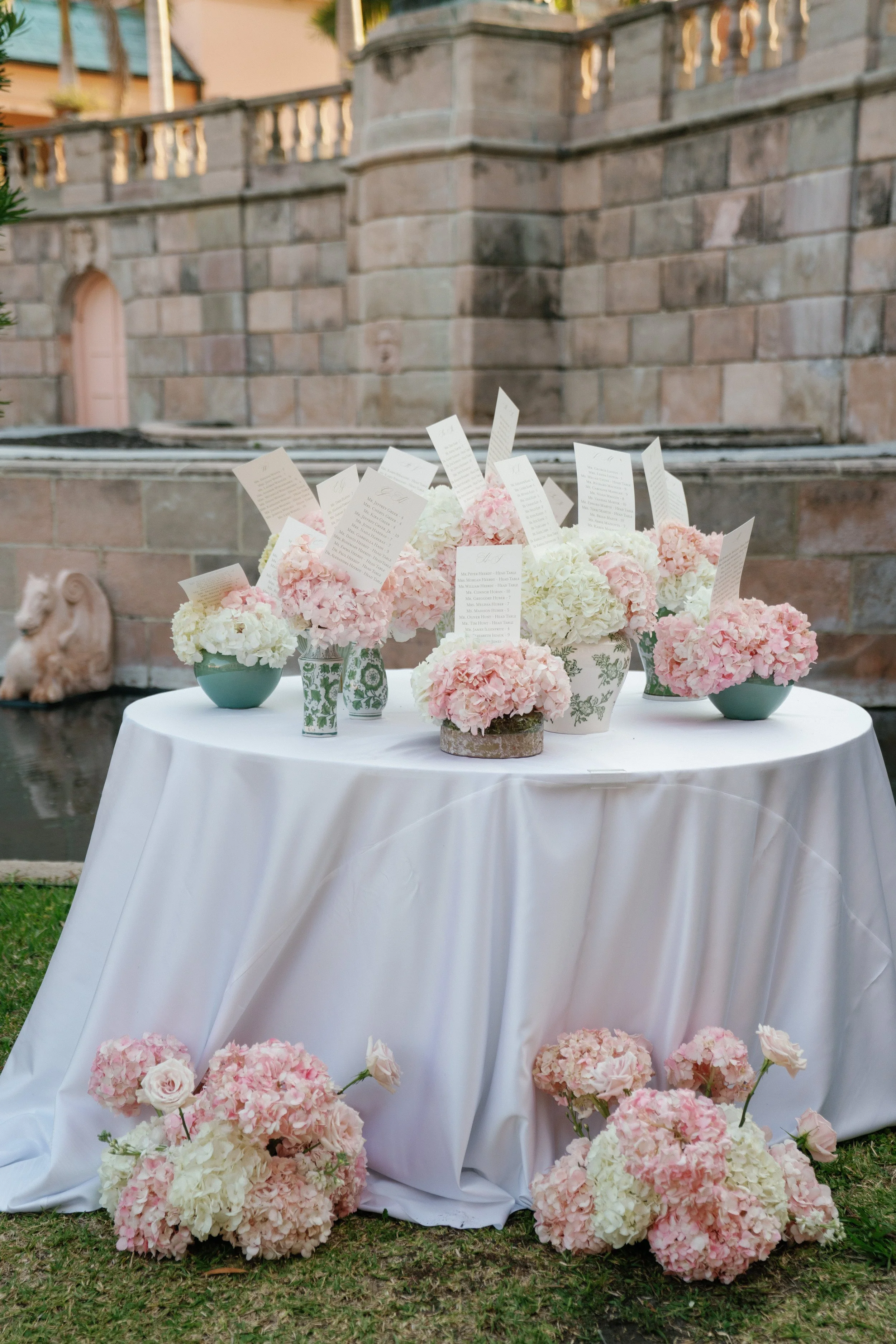 A round table with a white tablecloth decorated with pink and white hydrangea flower arrangements and scattered pink and white flowers on the ground in front, set outdoors with a stone wall and grass in the background, likely for a wedding or event.