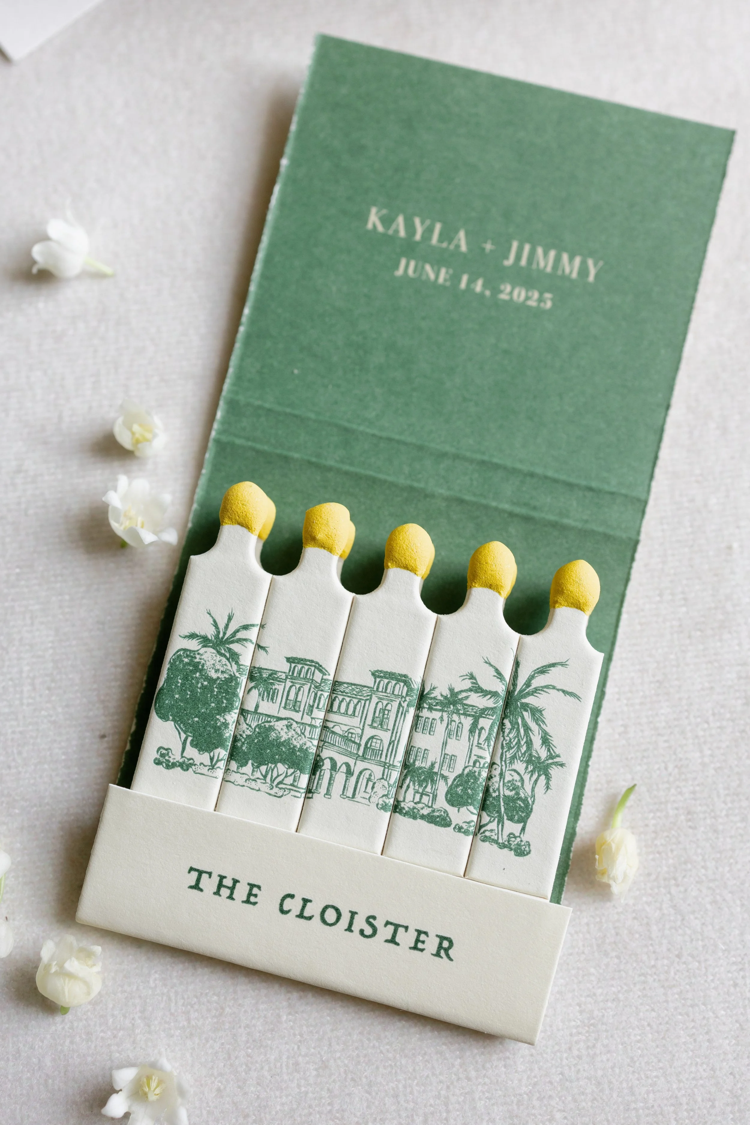 A matchbook with illustrations of a historic building, palm trees, and the text 'The Cloister' and 'The Cloister' printed on it. A green card with white text reads 'Kayla + Jimmy June 14, 2025'. Small white flowers and petals are scattered around on a light-colored surface.