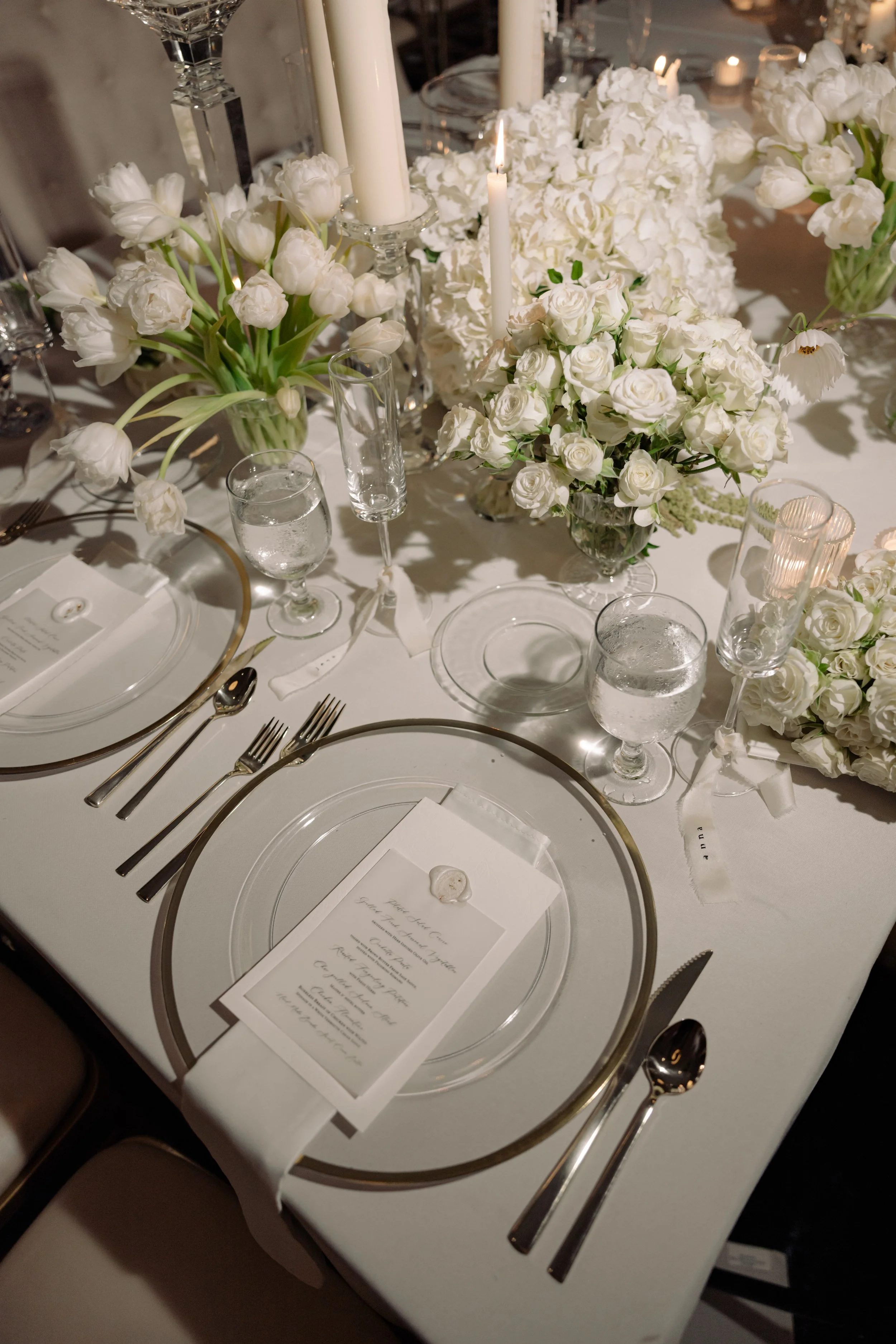 Elegant dining table decorated with white roses, tulips, candlesticks with tall candles, glasses, and a menu card, set for a formal event or celebration.