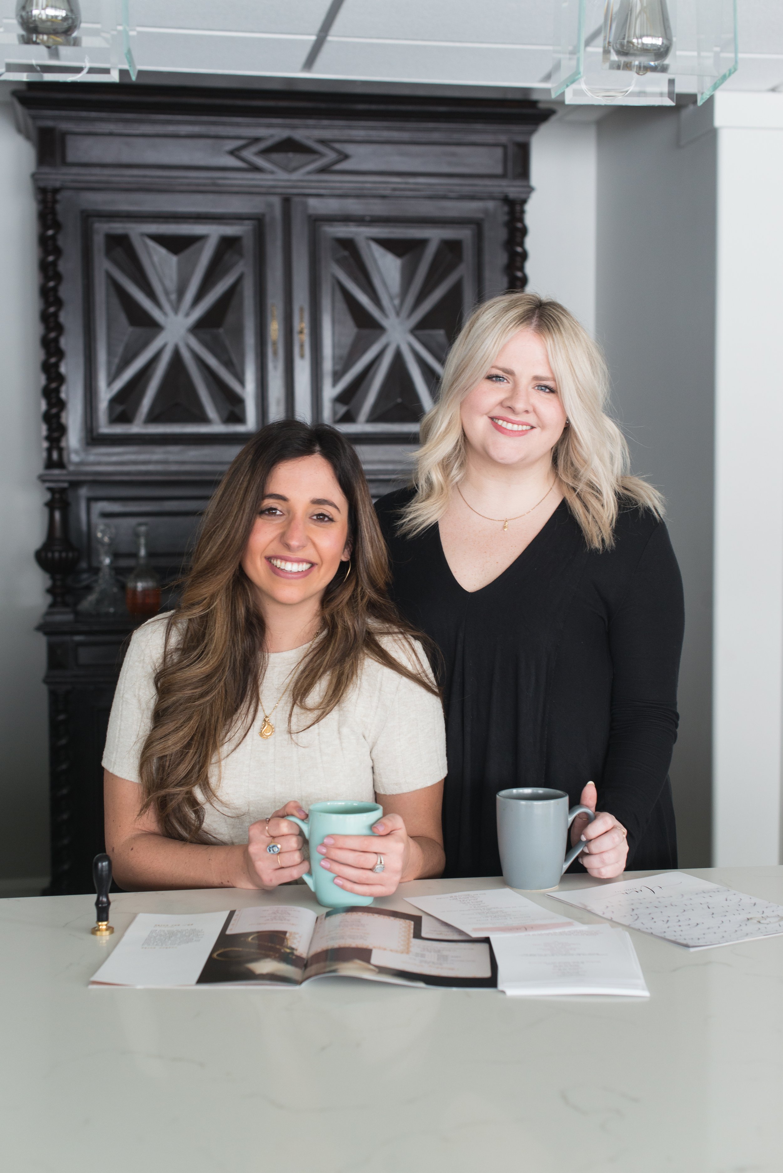 Two women smiling in a kitchen, sitting at a table with coffee mugs and open magazines.