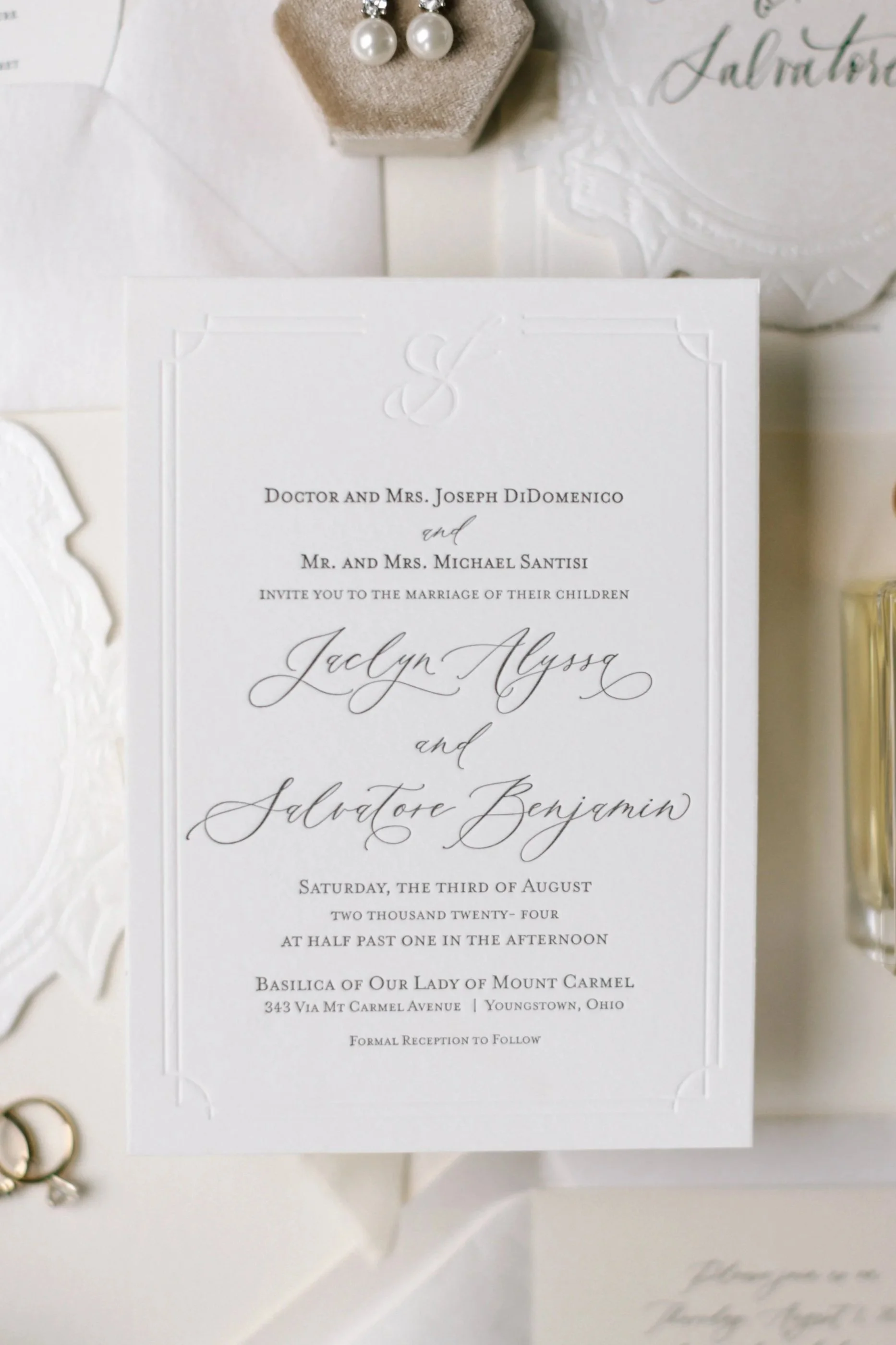 Wedding invitation with elegant black text on white paper, decorated with embossed designs, surrounded by jewelry and wedding accessories.