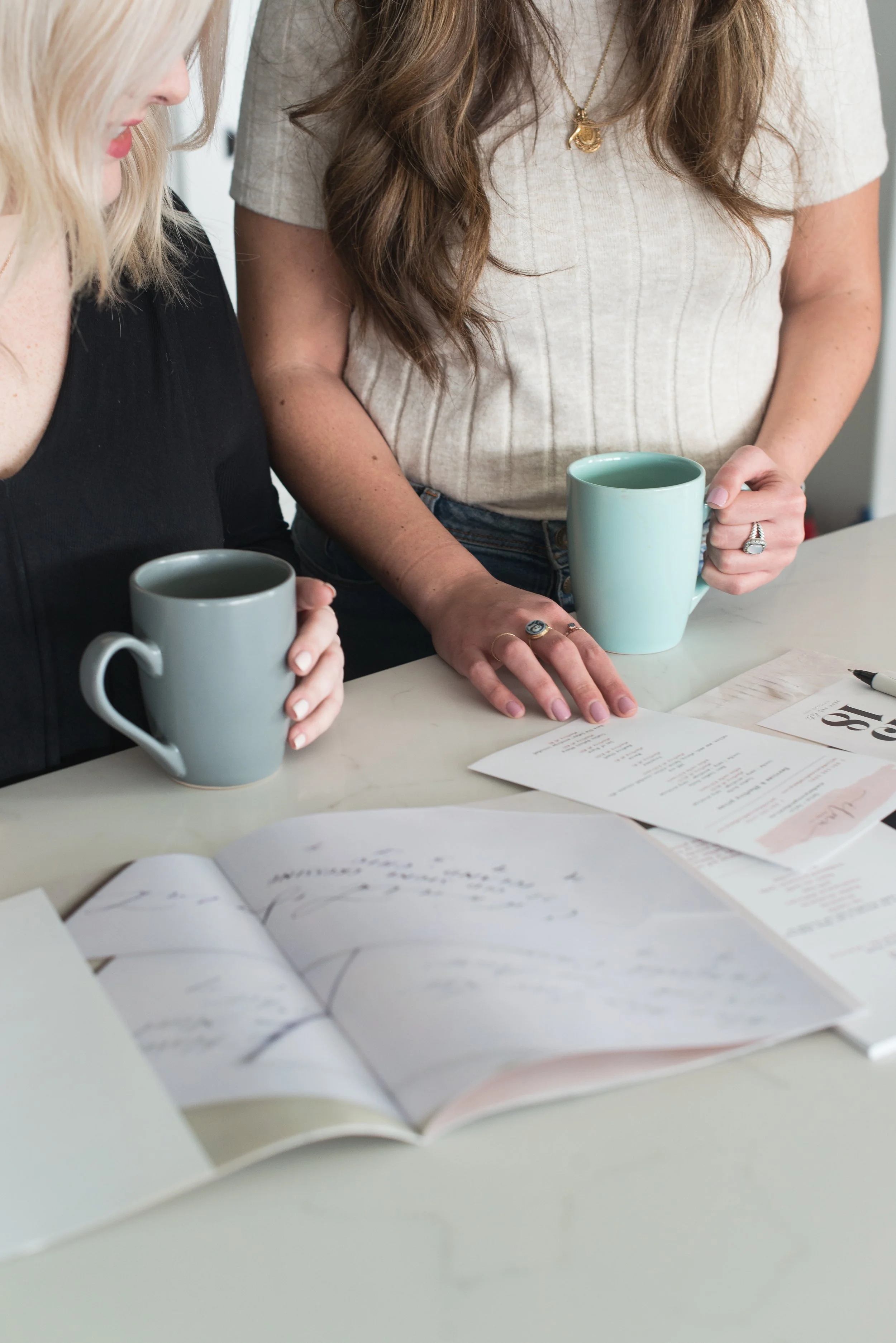 Two women holding light blue coffee mugs, sitting at a table with open notebooks, papers, and pens, engaged in a discussion or meeting.