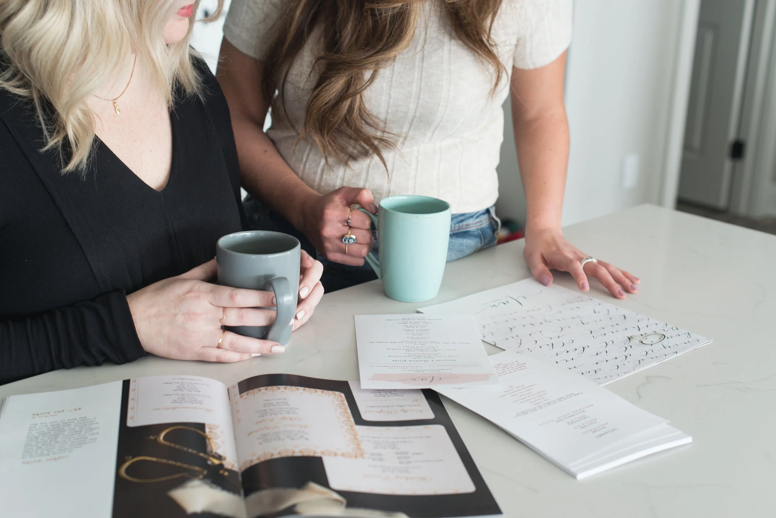 Two women standing at a table with coffee mugs and open booklets, one woman with blonde hair wearing a black top, the other with brown hair wearing a beige top, looking at handwritten calligraphy sheets.
