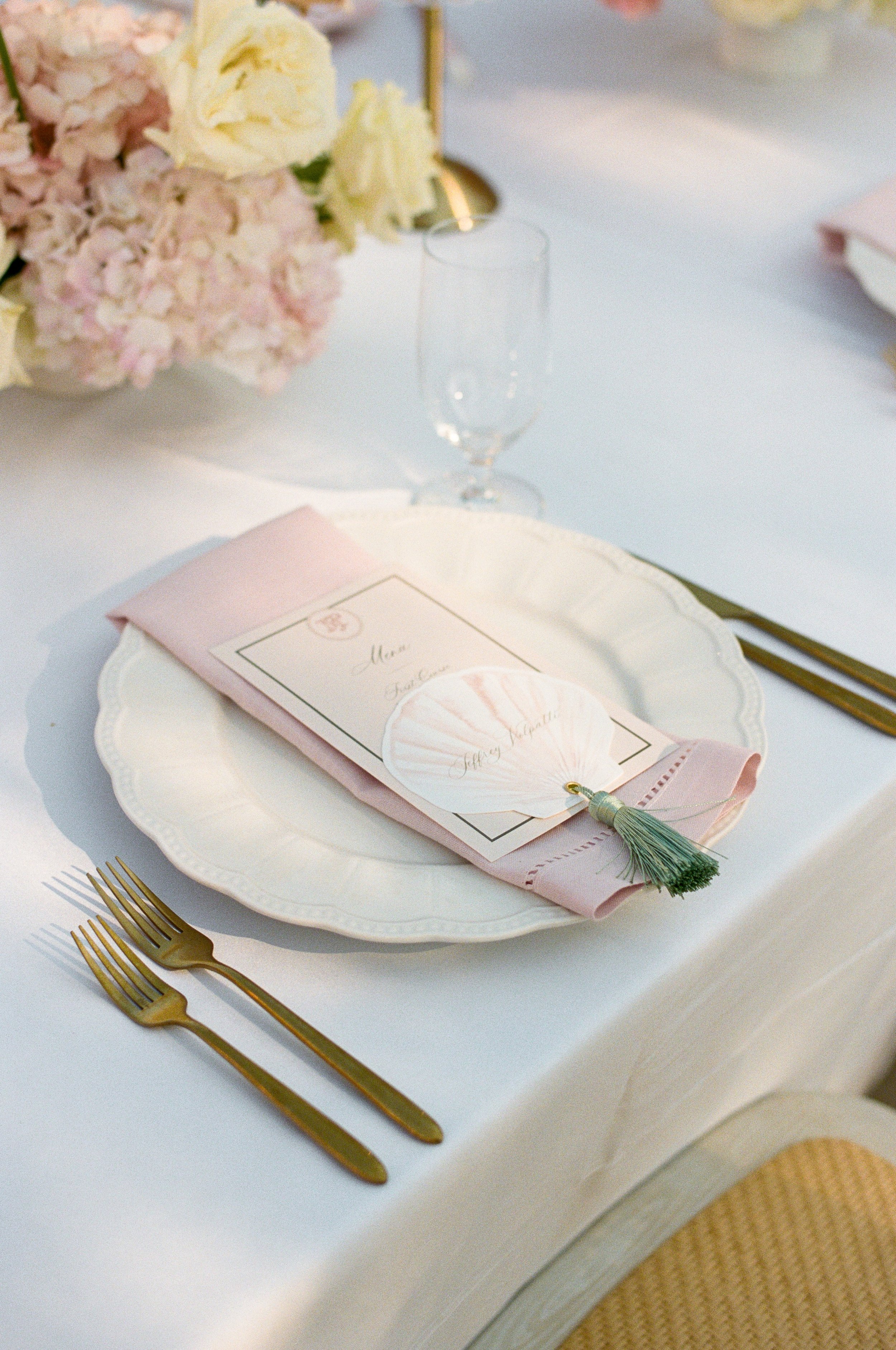A table setting with white plate, pink napkin, and a tassel, placed on a white tablecloth. In the background, there is a floral centerpiece with pink and white flowers and a glass of water.