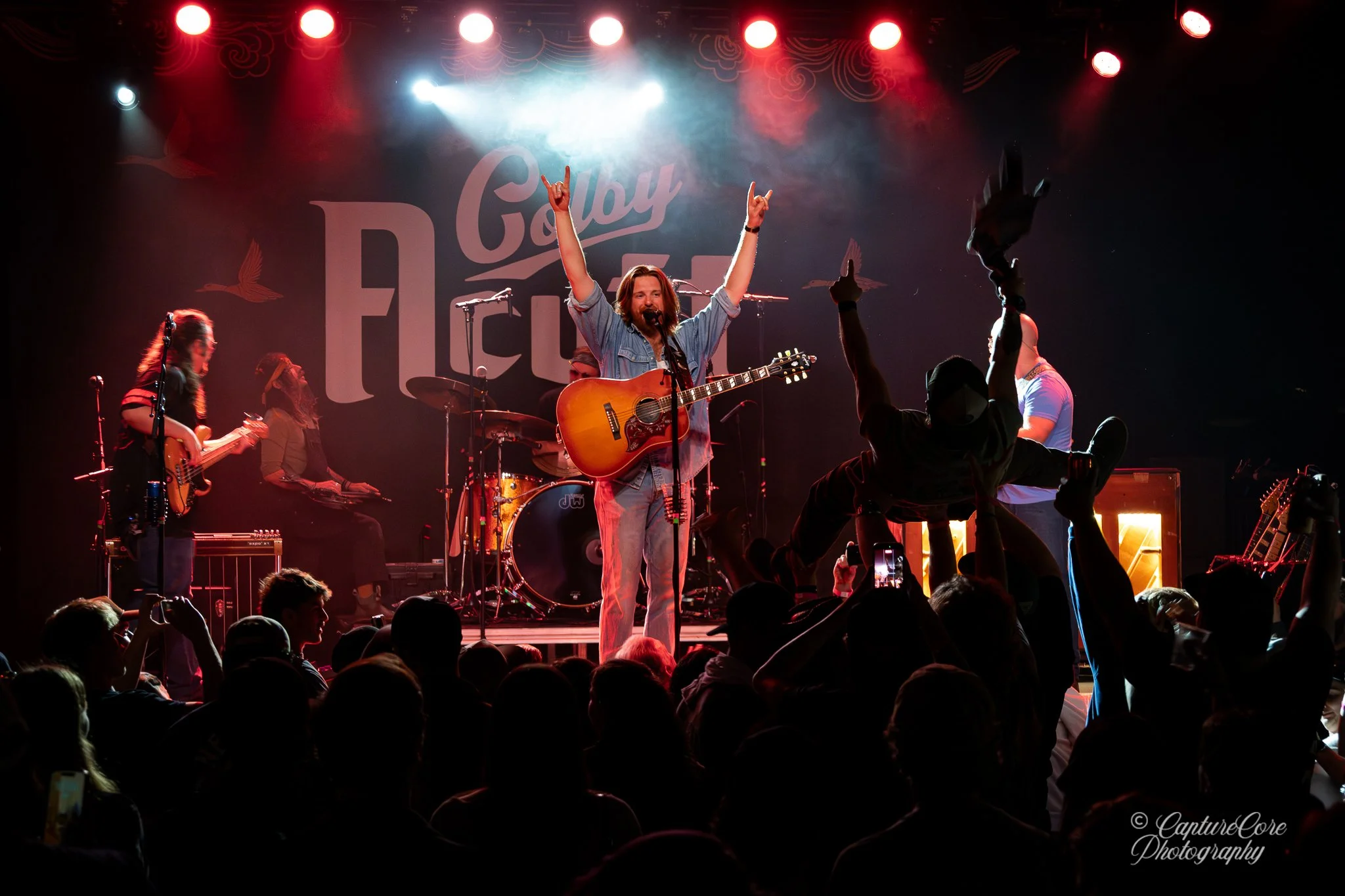 A band performs on stage with a lead singer playing guitar, raising both hands with peace signs, surrounded by band members playing guitar and drums, audience with raised hands in front, colorful stage lights, and a backdrop that says 'Gallery Fest'.