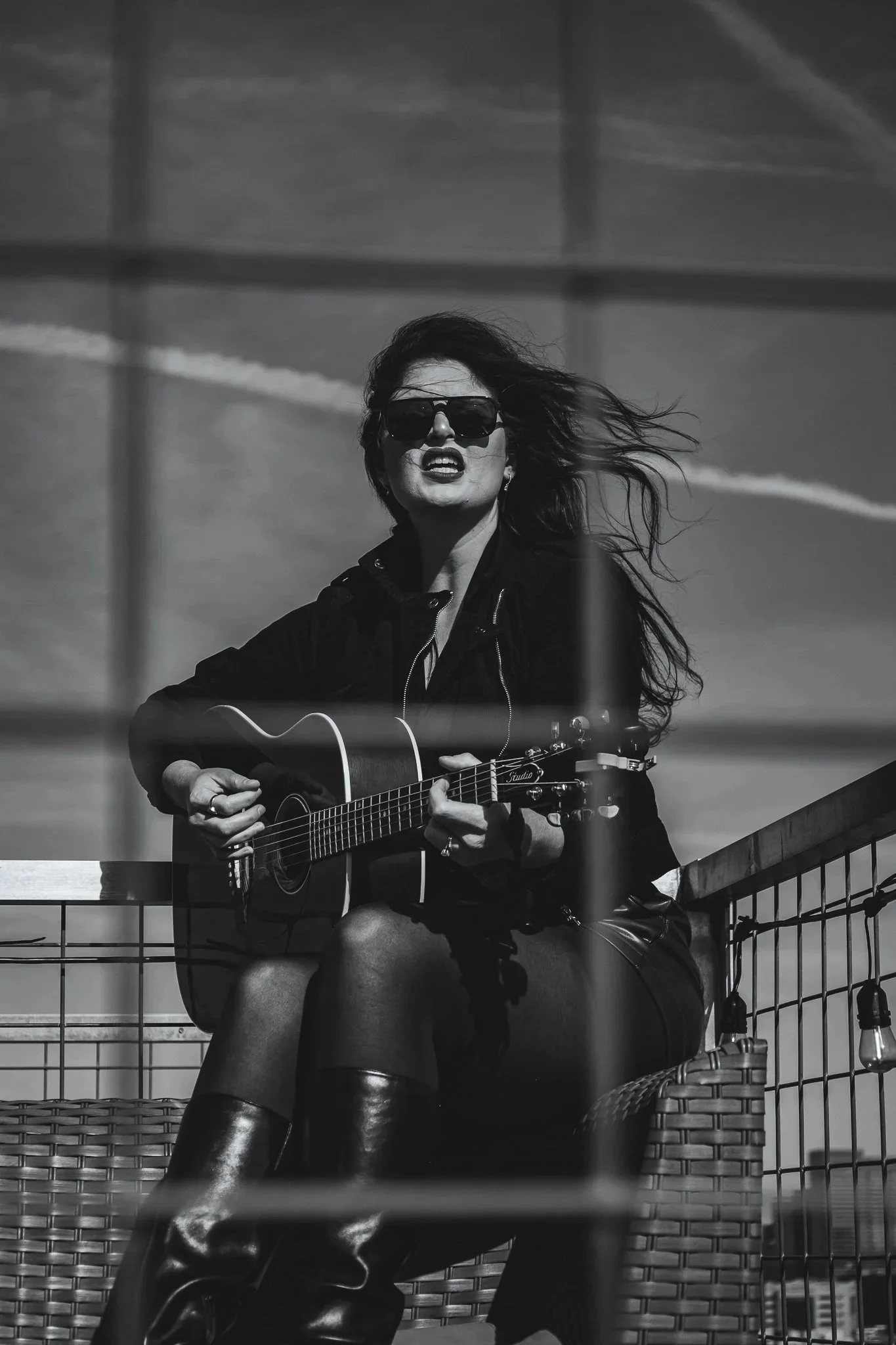 A woman with long hair wearing sunglasses and a dark jacket, playing an acoustic guitar while sitting outdoors on a wicker chair. There is a metal railing behind her and the sky in the background.