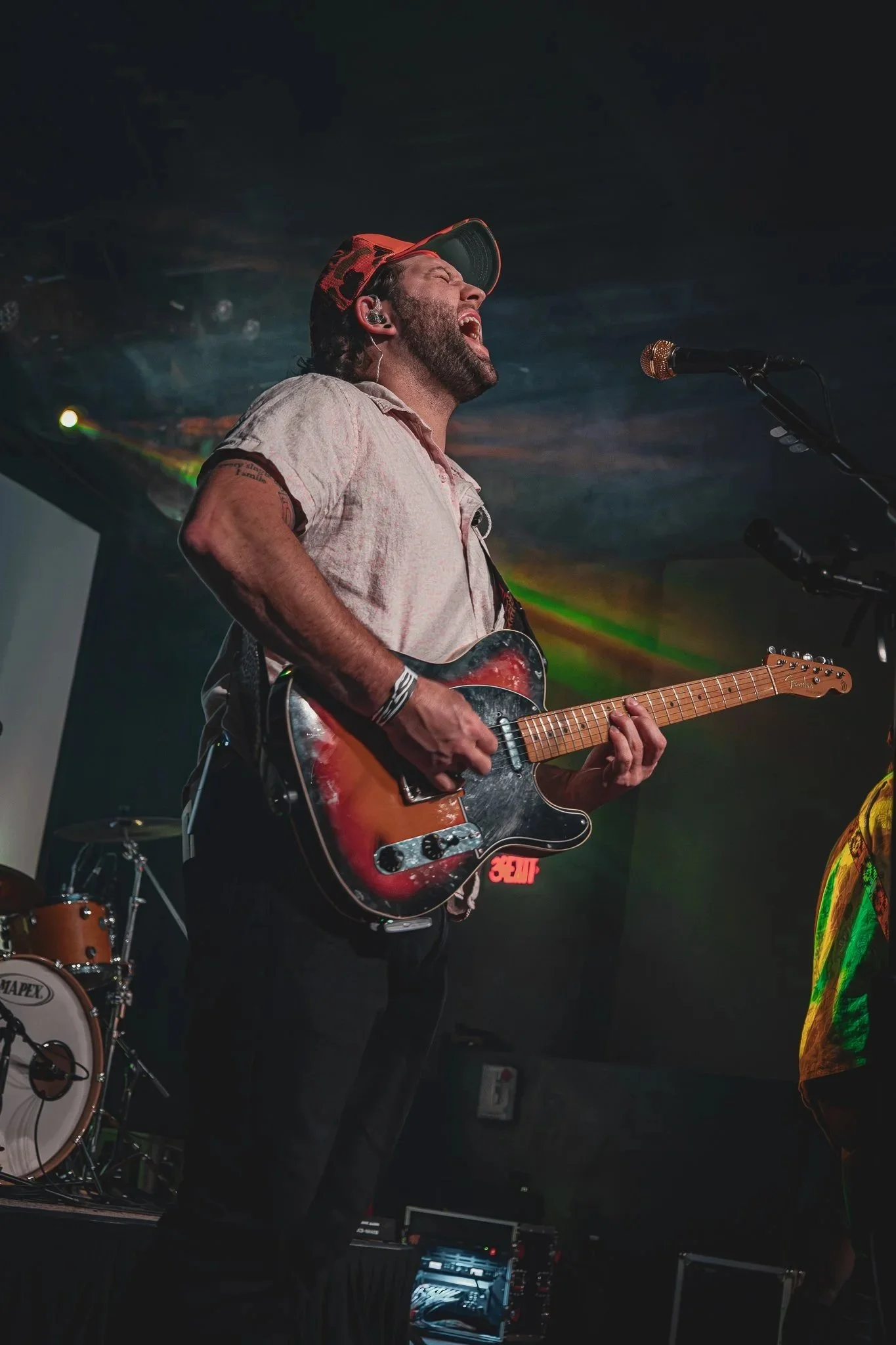 A man singing passionately while playing an electric guitar on stage, wearing a beige shirt, a baseball cap, and in-ear monitors, with a drum set and stage equipment visible in the background.