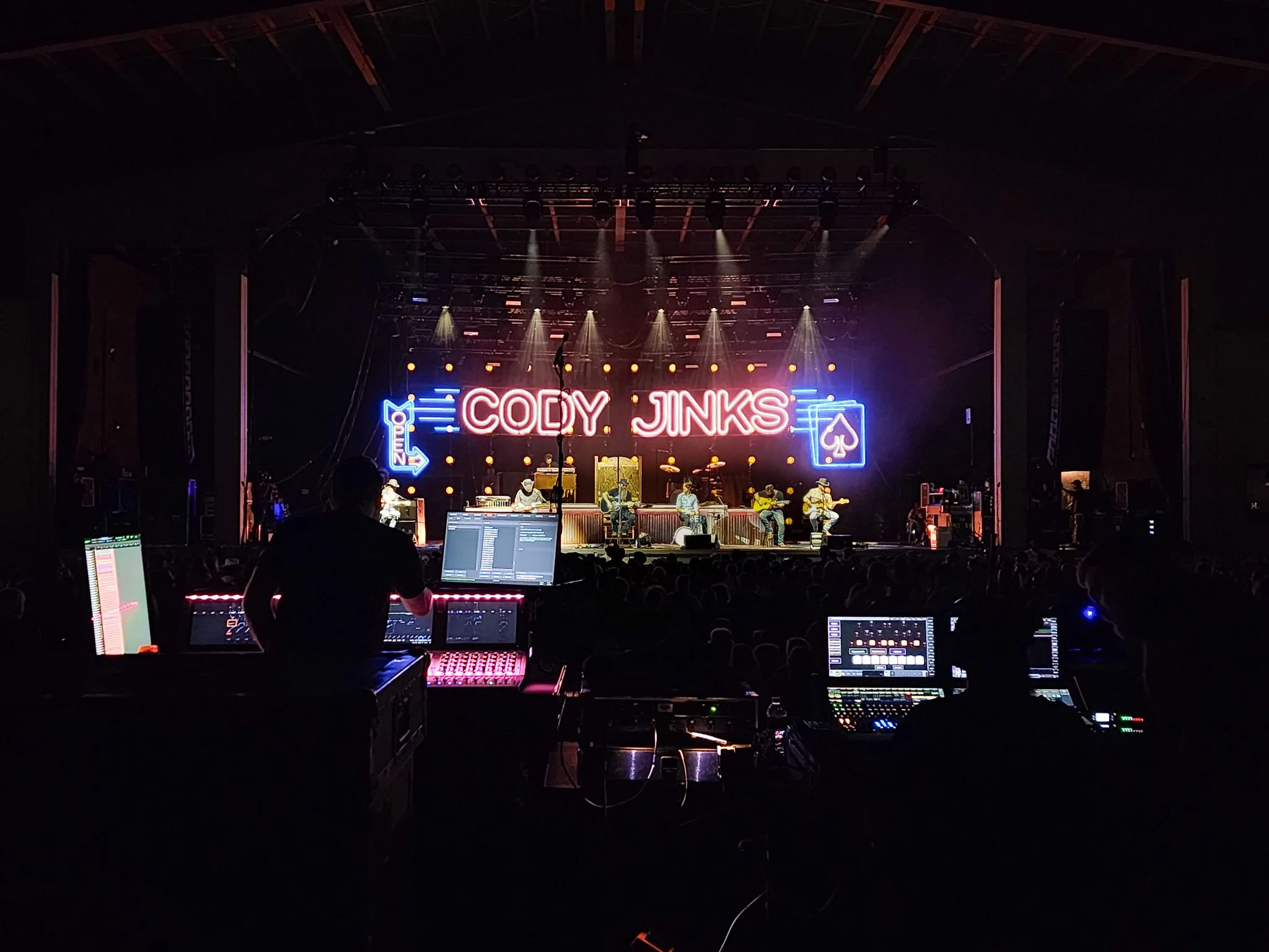 A stage with live music performance featuring neon signs displaying the name Cody Jinks, surrounded by stage lights and musical equipment, with a dark audience area in the foreground.