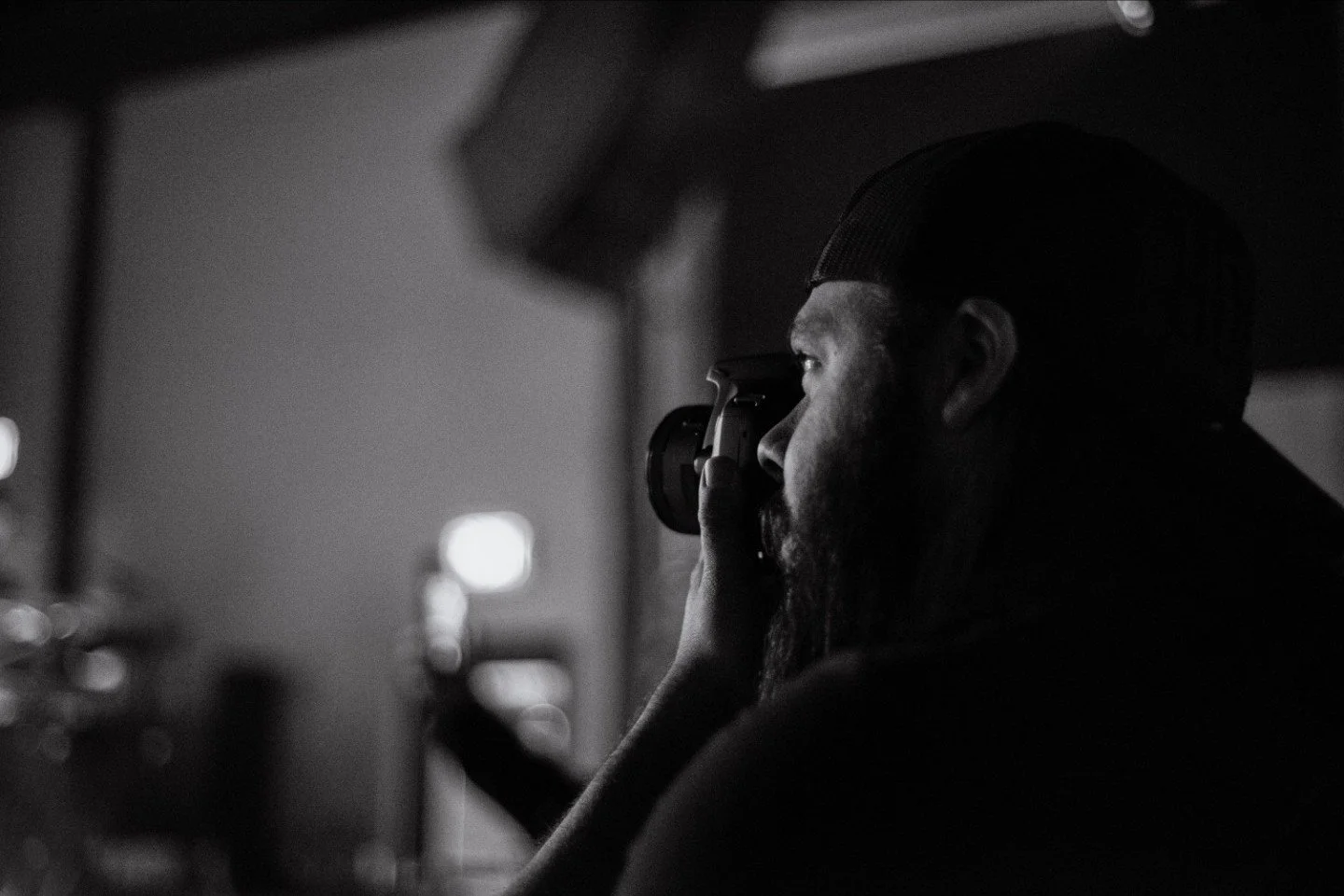 Black and white photo of a bearded man wearing a baseball cap, looking through a camera's viewfinder indoors.