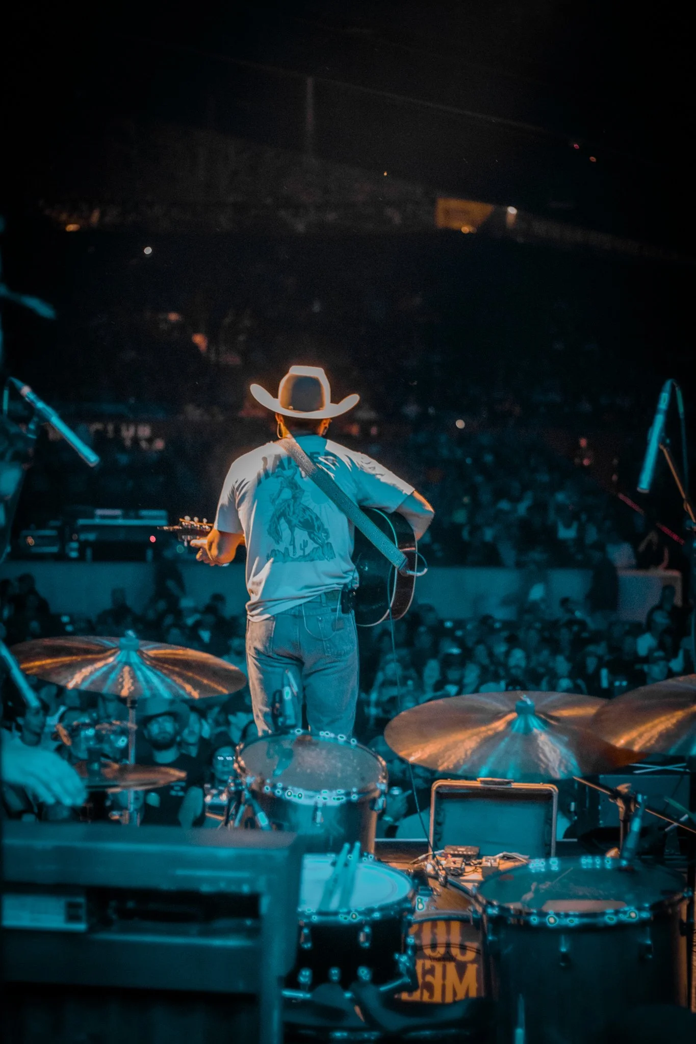 Musician with a cowboy hat playing guitar on stage during a concert, with drums and audience in the background.