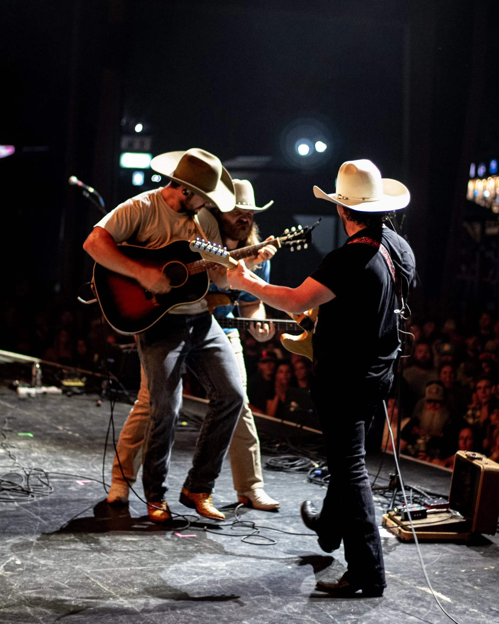 Three musicians wearing cowboy hats perform on a stage with guitars and microphones, with an audience in the background.