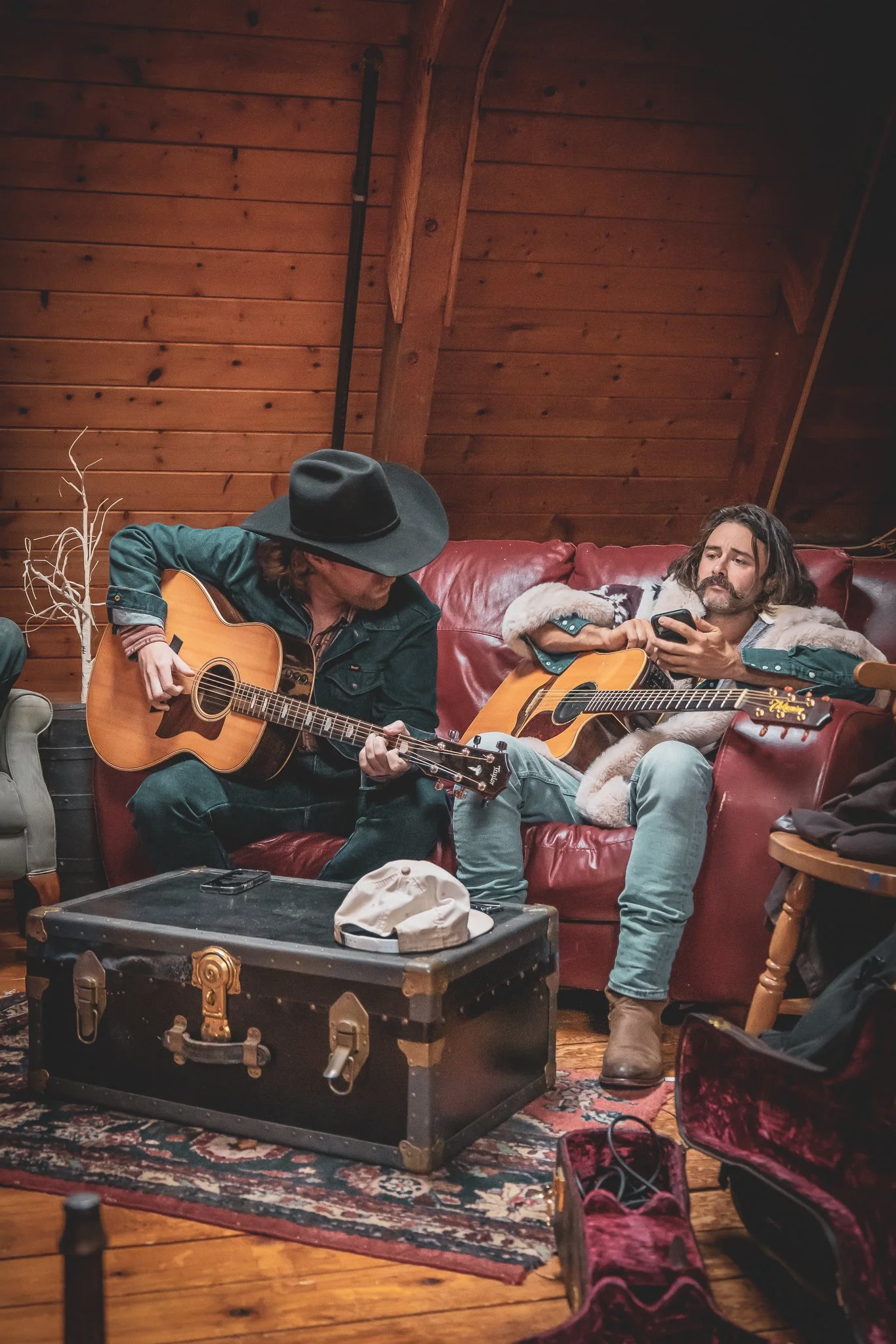Two men with guitars sitting on a red leather couch in a wooden room, one wearing a black hat and a denim jacket, the other with long hair and a fur-lined coat, looking at a phone. There is a trunk and a stroller with pink shoes nearby.