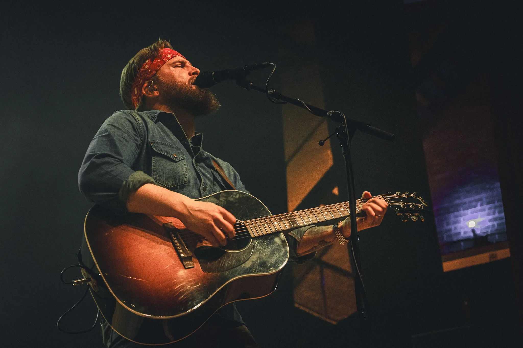 A man with a red headband and beard singing into a microphone while playing an acoustic guitar on stage.