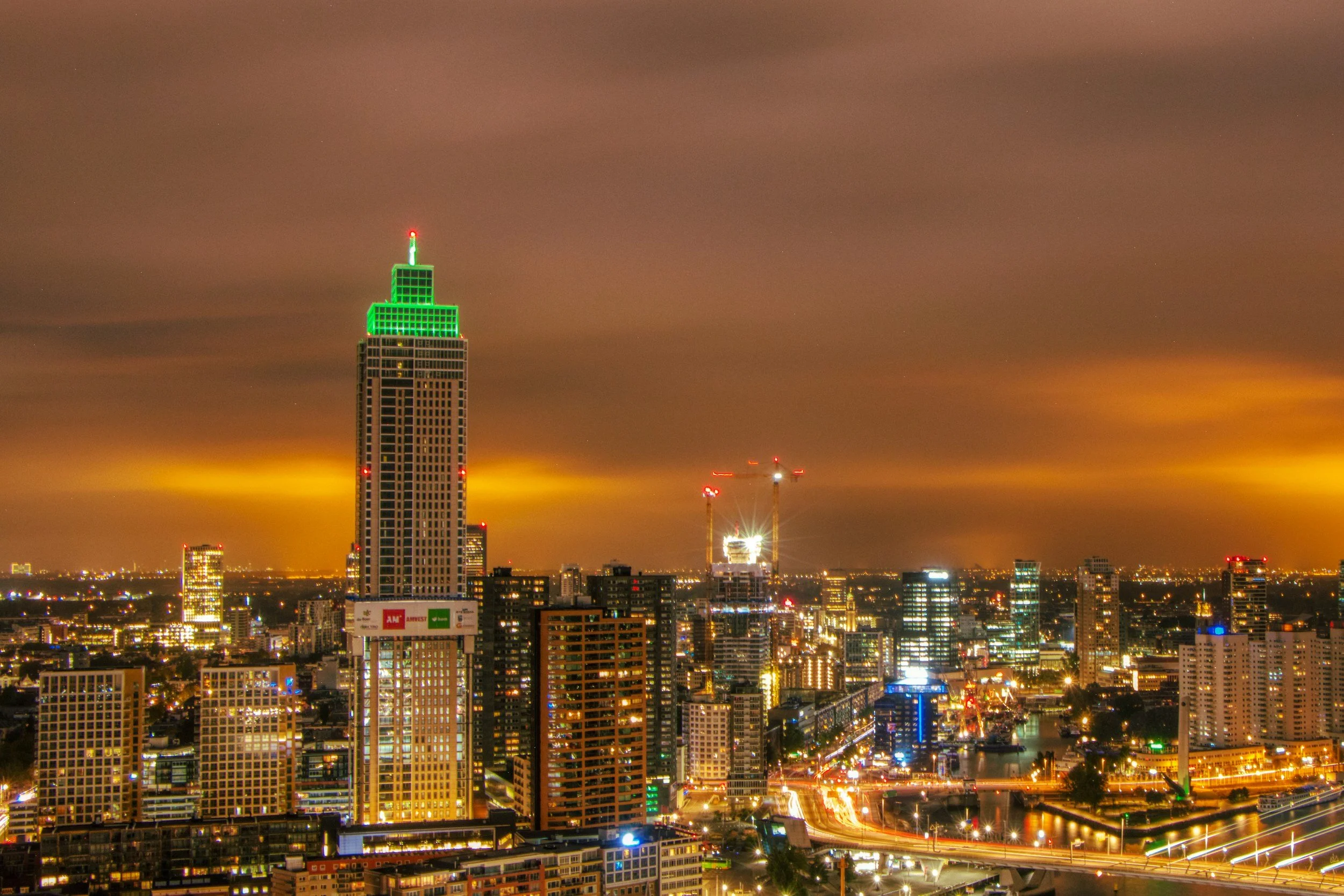 Uitzicht op de skyline van een grote stad 's avonds met een toren die groen verlicht is en een bewolkte hemel op de achtergrond.