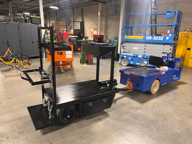 Industrial warehouse with various equipment, including a black cart with hooks and chains, a blue scissor lift, and yellow machinery, on a concrete floor.