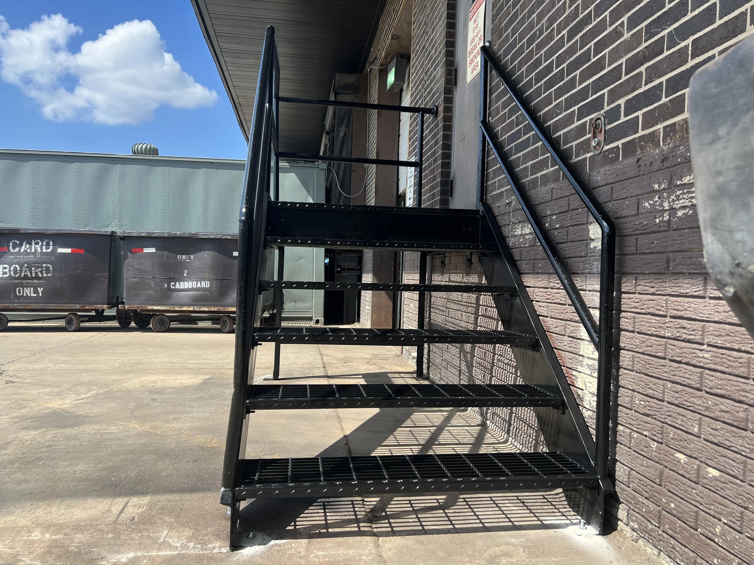 Black metal staircase or fire escape on a brick building exterior, with a blue sky and white clouds in the background.