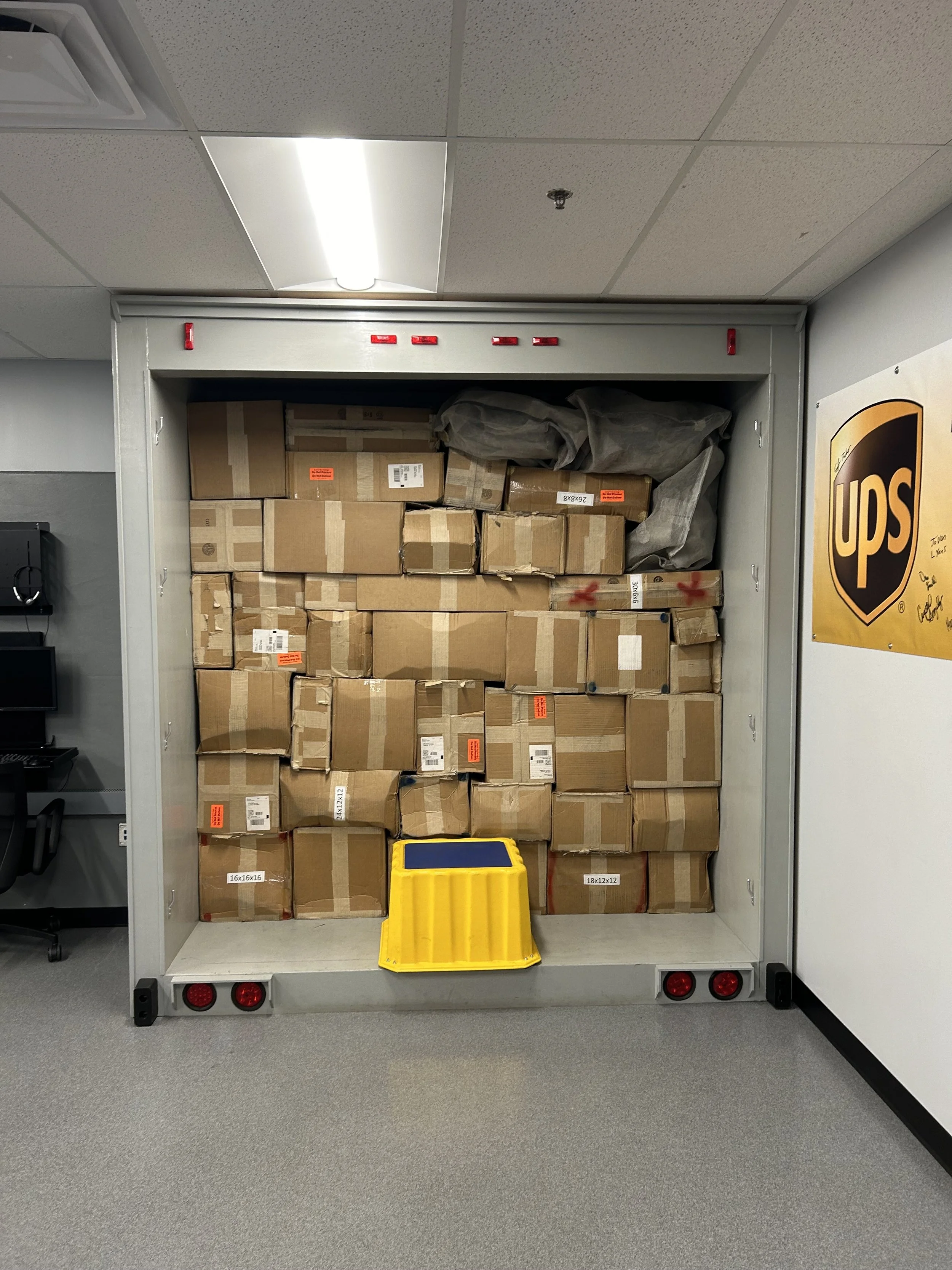 A UPS delivery truck loaded with stacked cardboard boxes and gray packaging material, with a yellow plastic container at the bottom. The truck has red reflectors and is in an indoor setting with gray carpeted flooring and fluorescent ceiling lights.