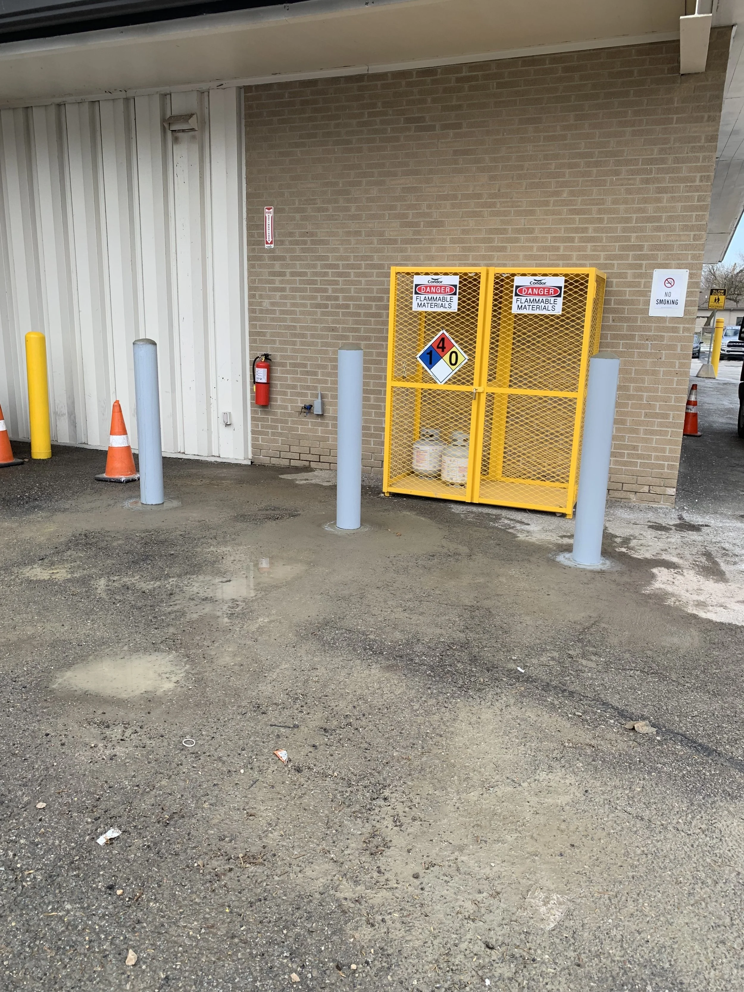 Image of a parking lot area with a brick wall, a yellow hazardous materials cage, a fire extinguisher, and traffic cones and bollards, with signs indicating danger and no smoking.