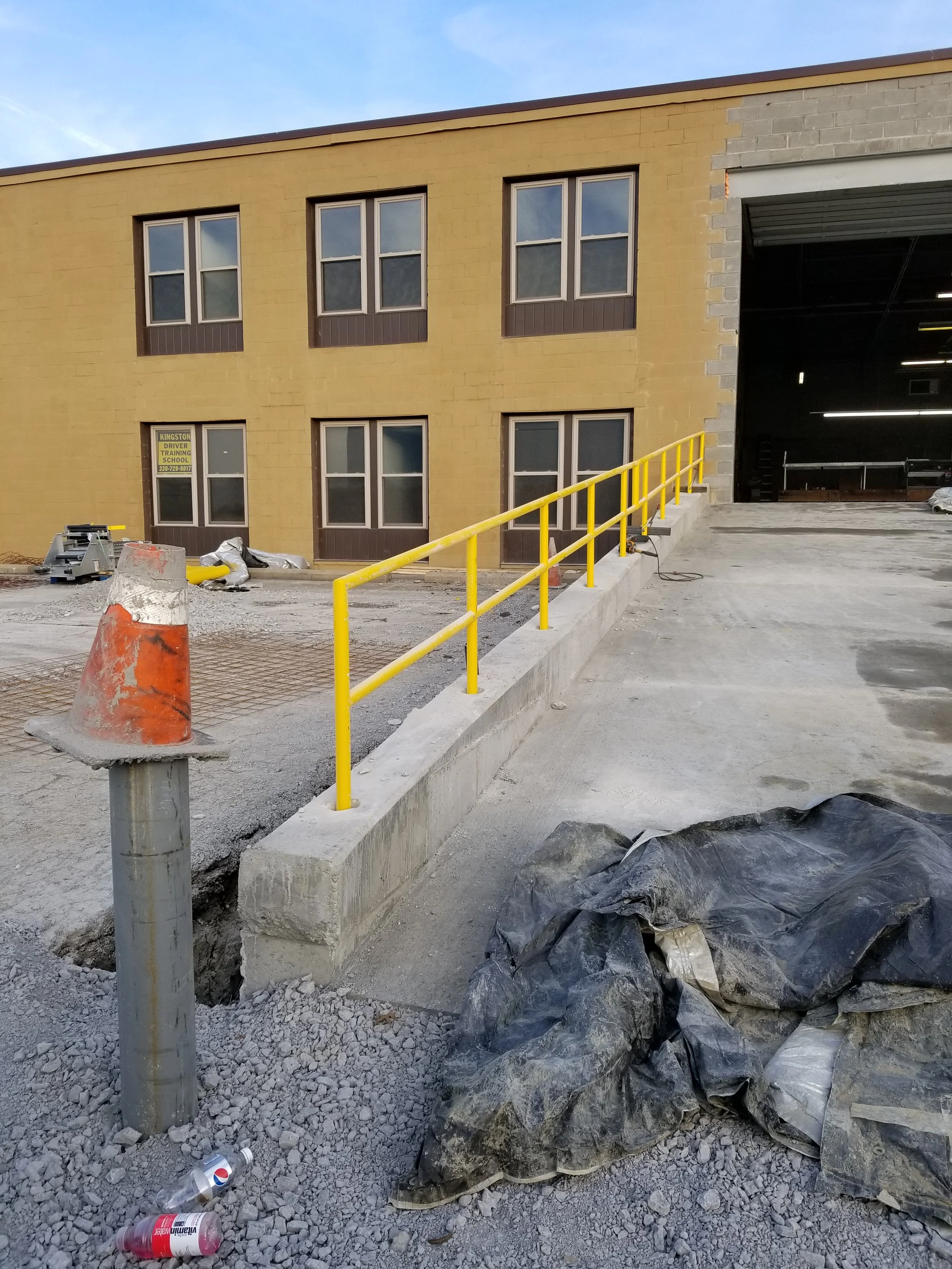 Construction site with yellow safety railing, gravel ground, construction debris, and a partially built yellow brick building with windows, an open garage door, and a blue sky.