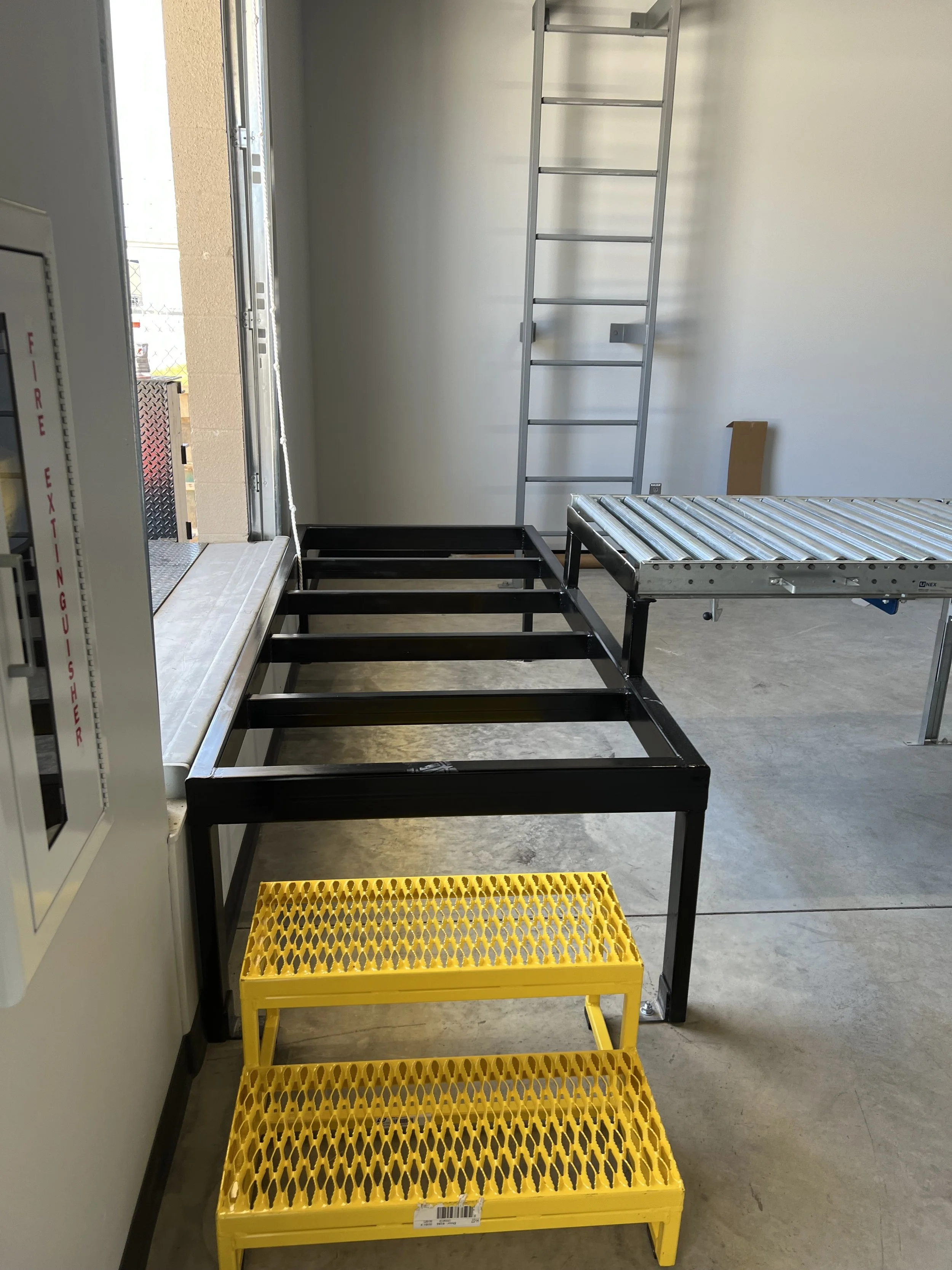 Empty metal bed frame with black finish, yellow plastic step stool, and metal ladder in a room near a door with a fire exit cabinet on the left.
