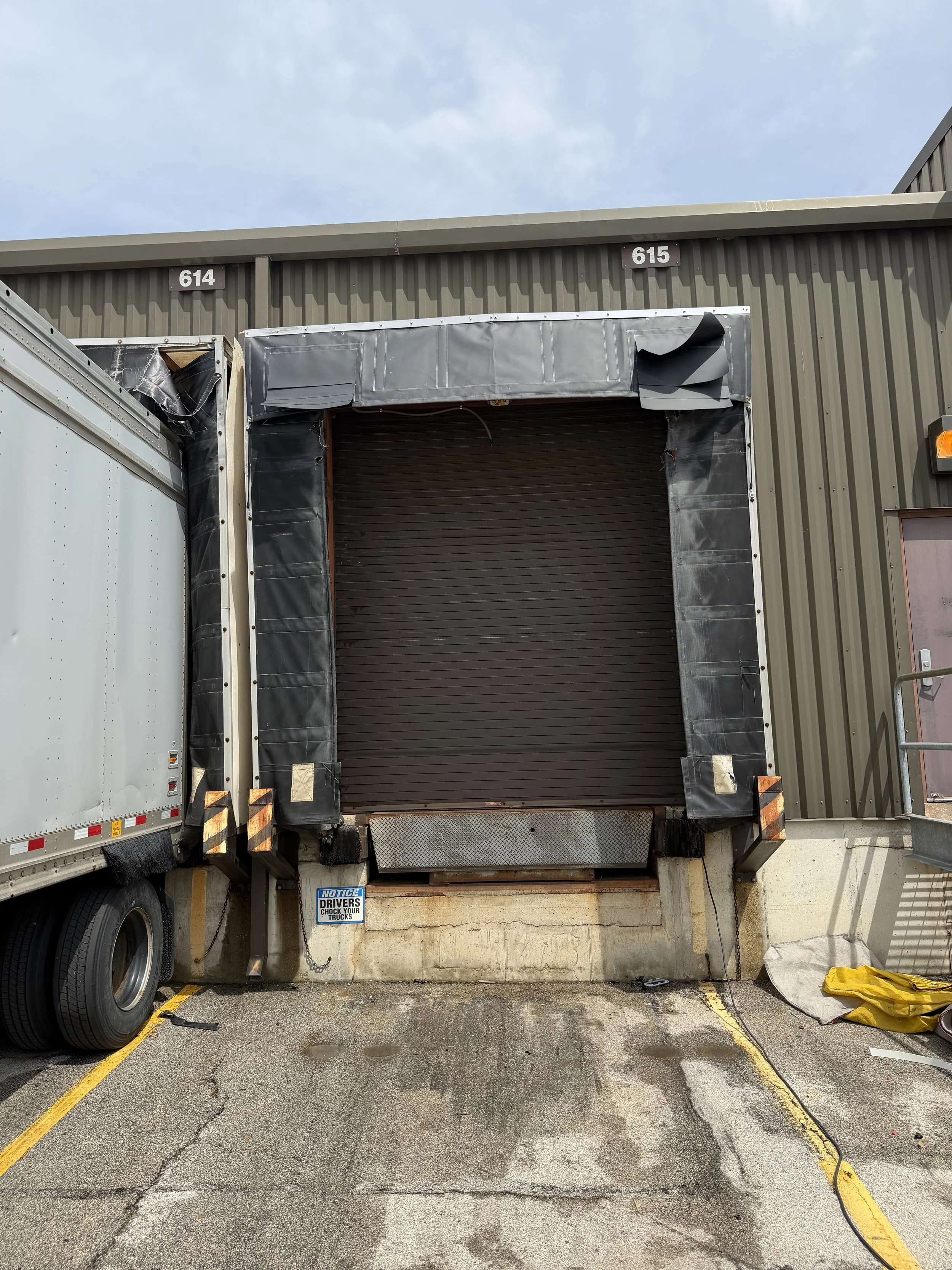 Loading dock with a partially open roll-up door on a warehouse building. Two trucks are parked in front, one on each side. The parking area has yellow painted lines and a sign warning drivers about checking trucks.
