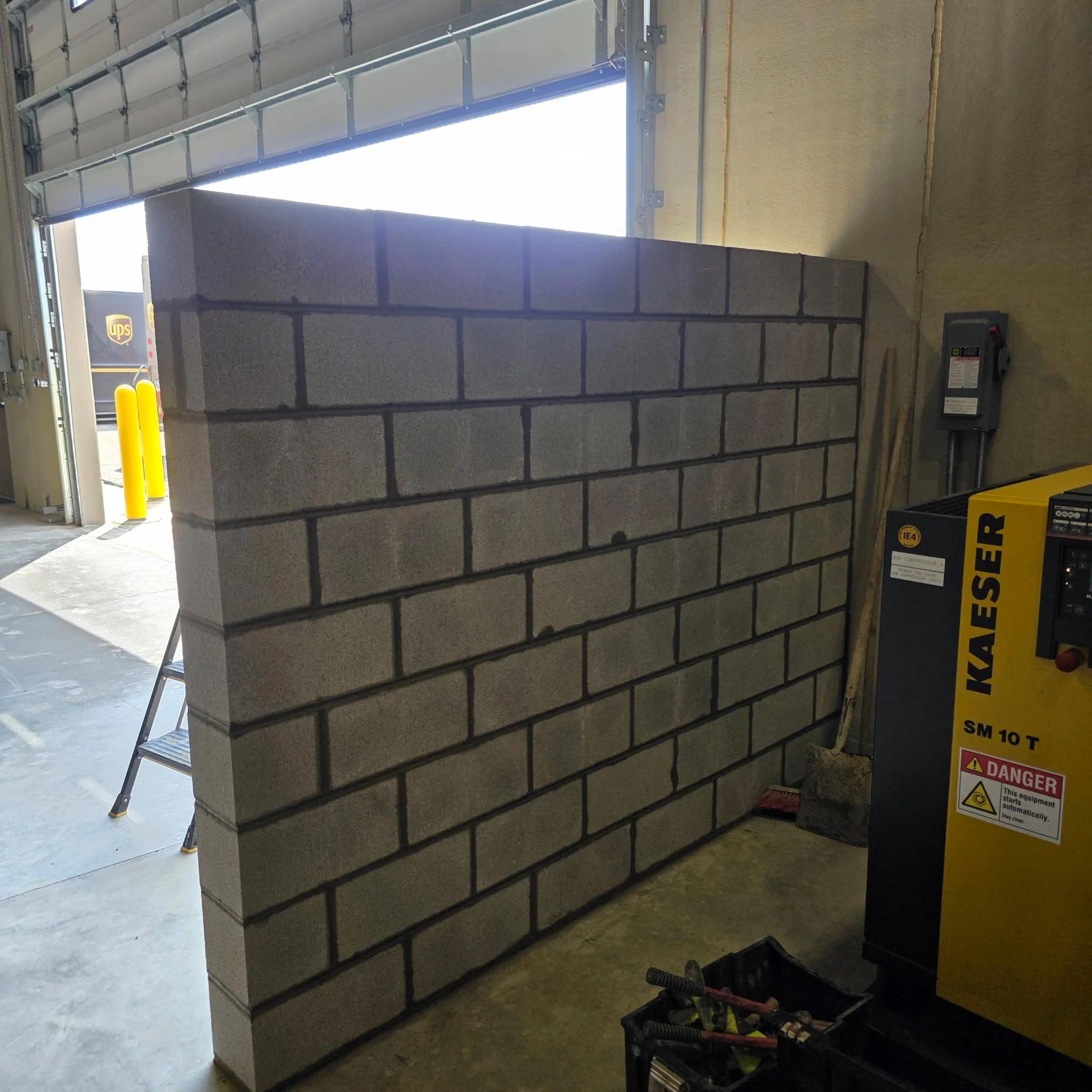 Interior of a garage with a partially built cinder block wall. there is a gray wall on the right side with a yellow and black warning sign for air compressor equipment. There is a ladder leaning against the wall, and some tools and equipment on the floor and in a black crate. An opened garage door shows a delivery truck outside.