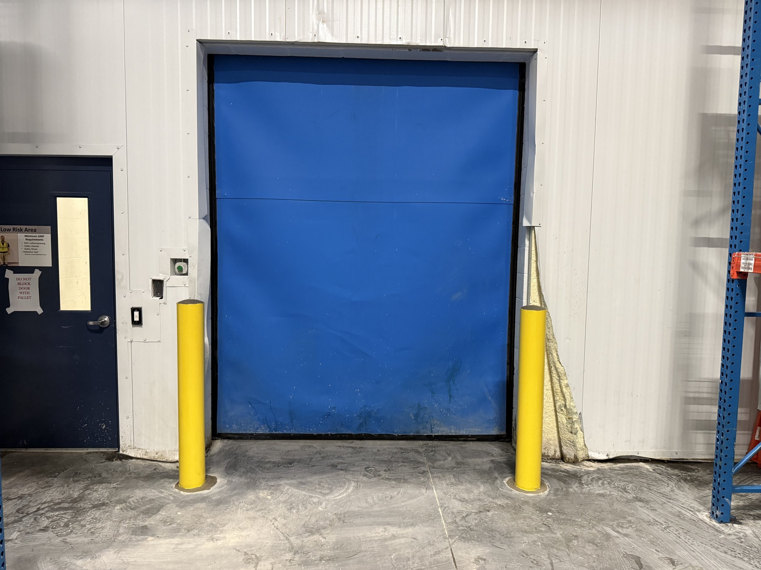 A large blue rolling door with two yellow safety bollards in front of it, situated in an industrial warehouse or storage facility.