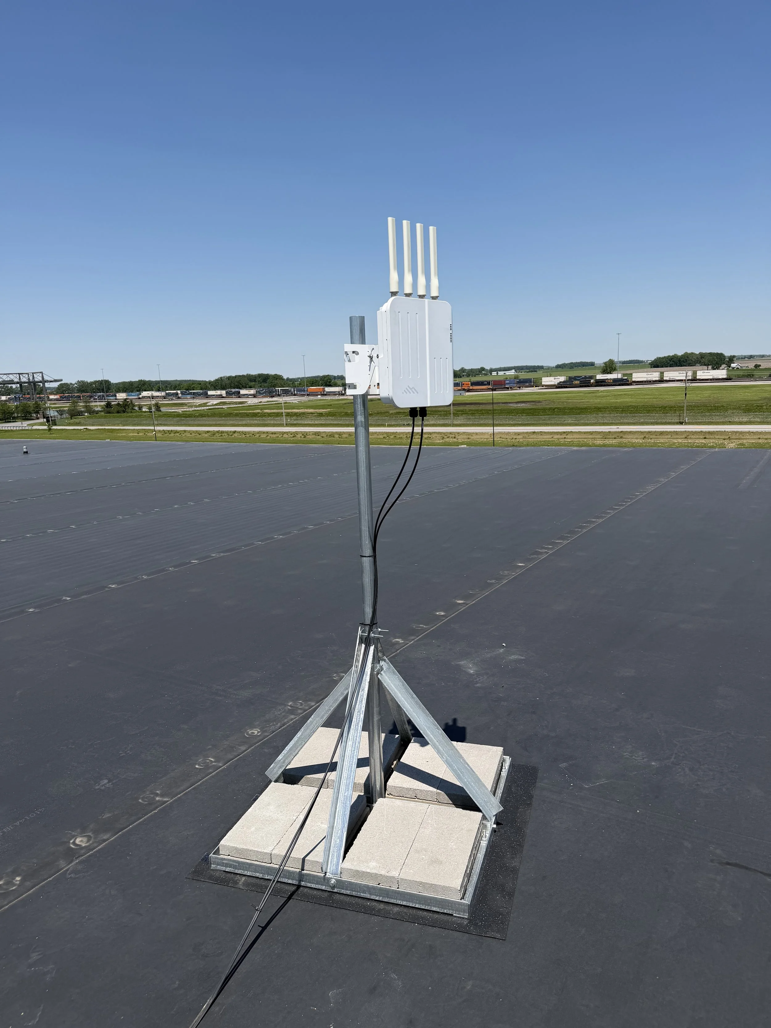 A wireless antenna mounted on a building's rooftop with a scenic background of train tracks, trains, trees, and a blue sky.