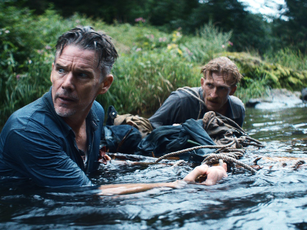 Two men are in a river surrounded by lush greenery, with some camping gear and ropes in the water as they appear to be stranded or stranded.