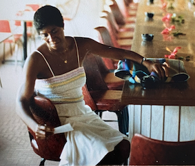 Woman in white dress sitting at a bar counter with a blue-colored drink and sunglasses on a sunny day