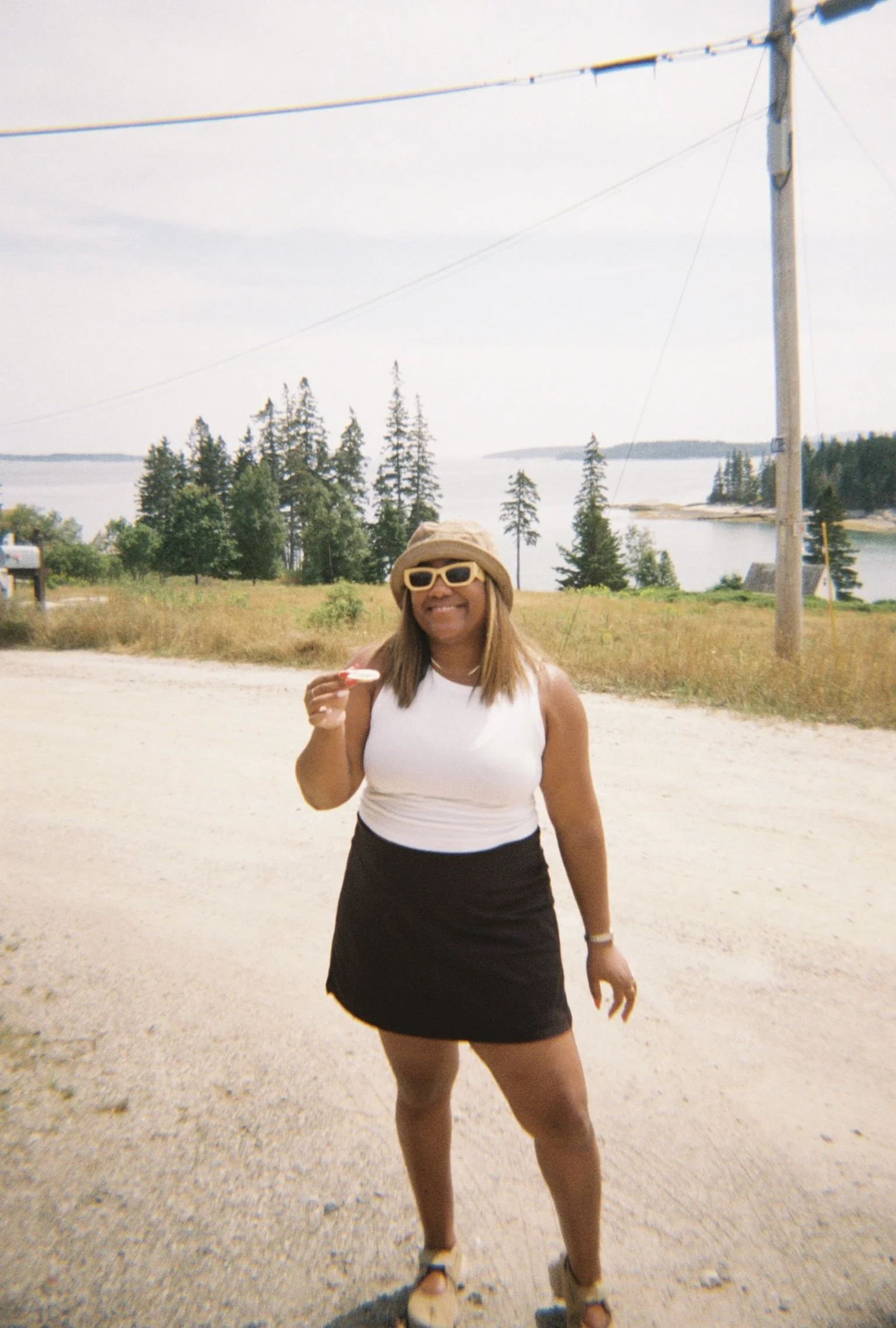 A woman wearing a beige sunhat, yellow sunglasses, a white sleeveless top, black shorts, and sandals standing outdoors near a body of water with trees and a utility pole in the background, smiling and holding an object.