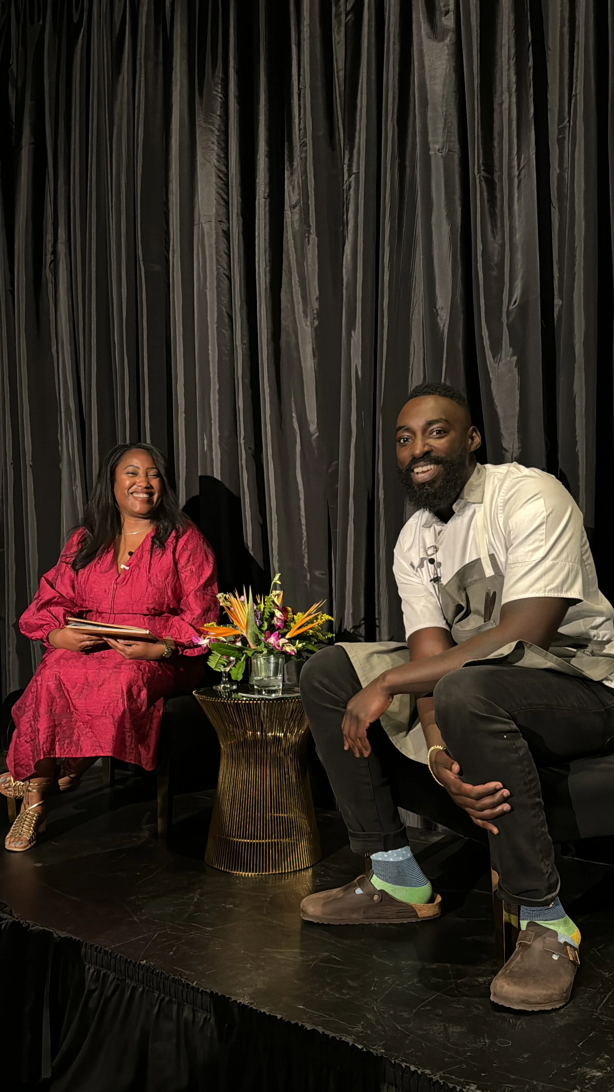 Quayla and Chef Eric Adjepong sitting on a stage with a black curtain in the background, a small table with a flower arrangement, both smiling. Quayla wearing a pink dress and Chef Eric in casual beige and black clothing.