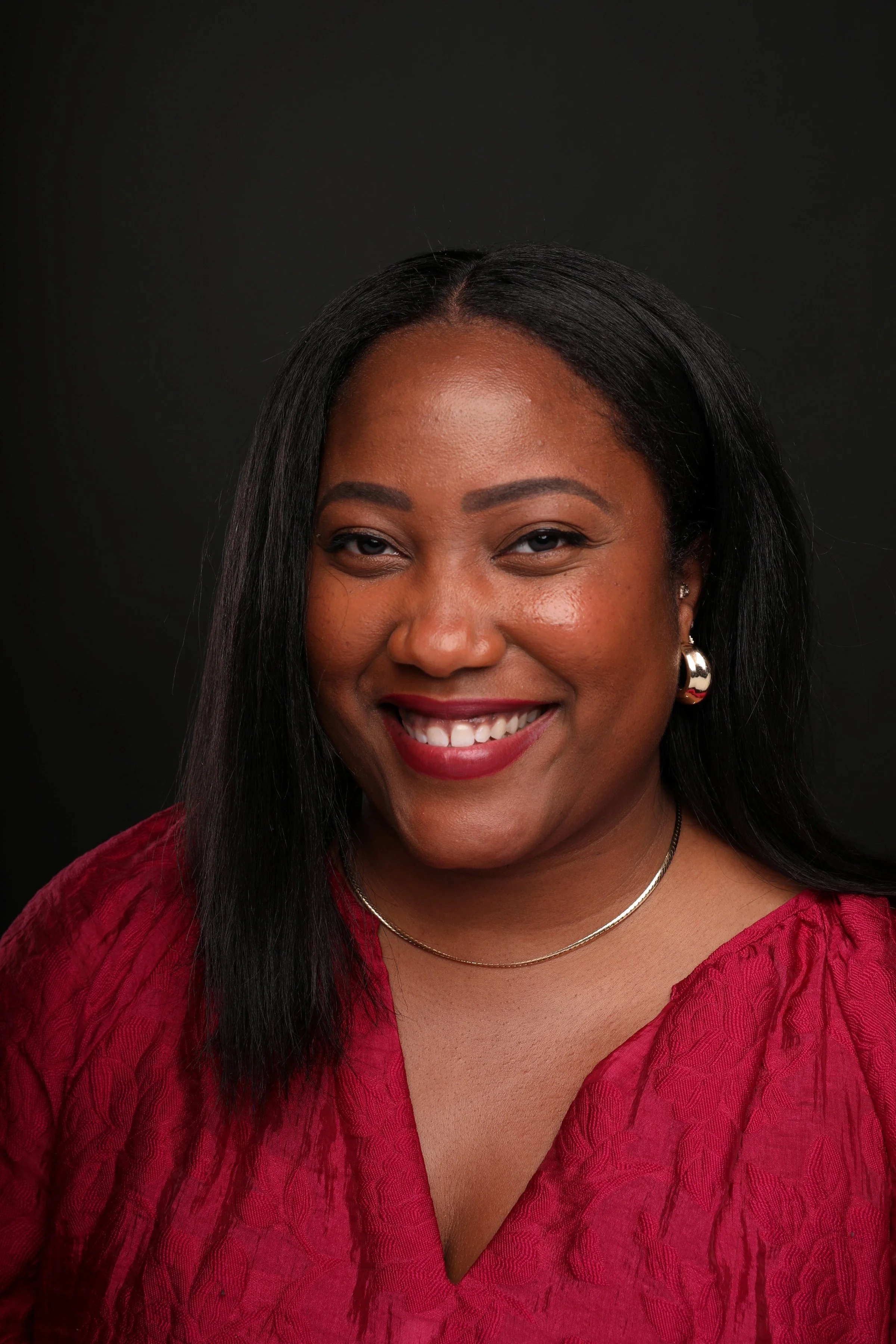 A smiling woman with black hair, wearing earrings, a gold necklace, and a red top, against a black background.