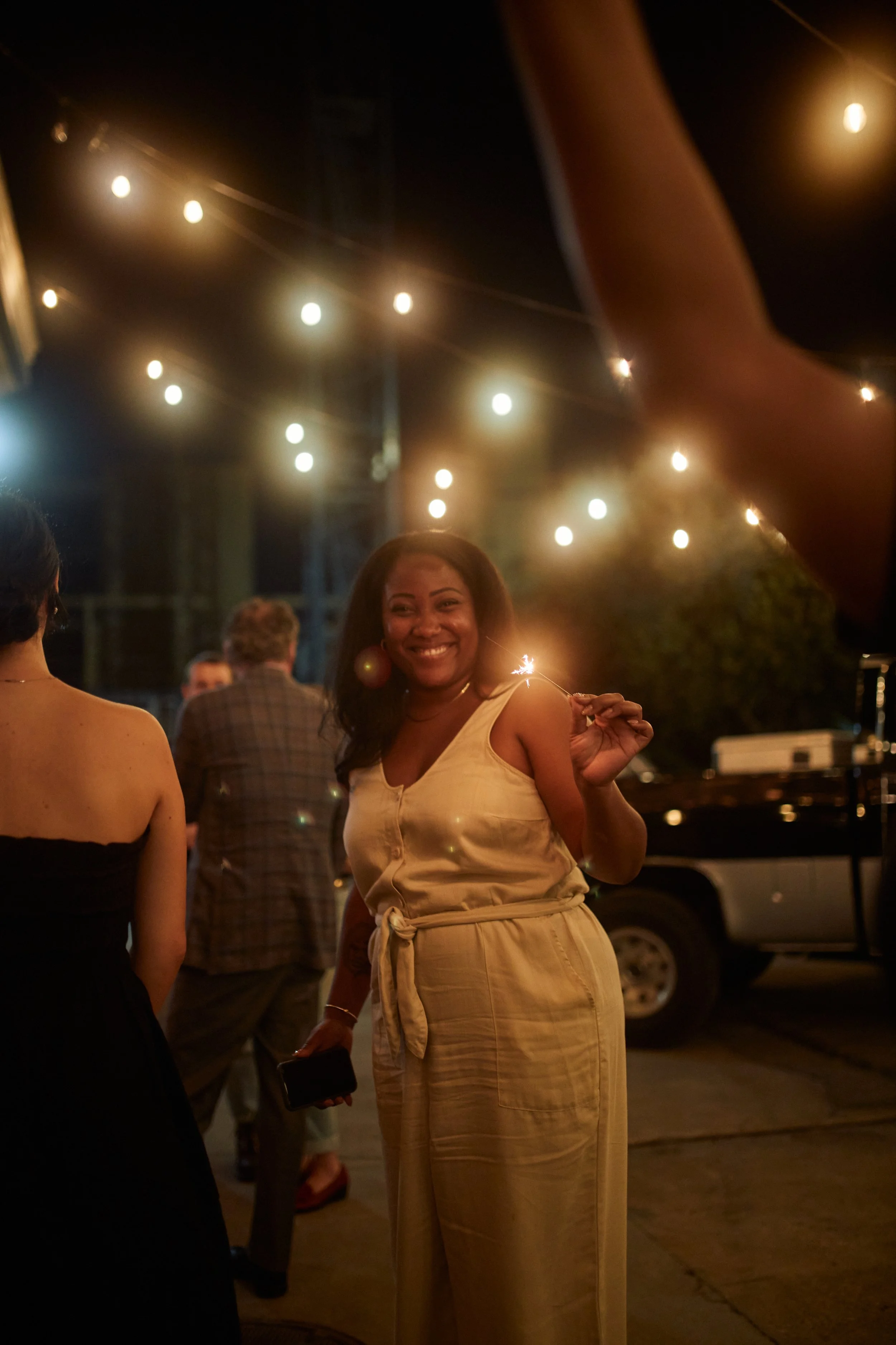 A woman smiling at night, holding a sparkler, standing among other people with string lights overhead.