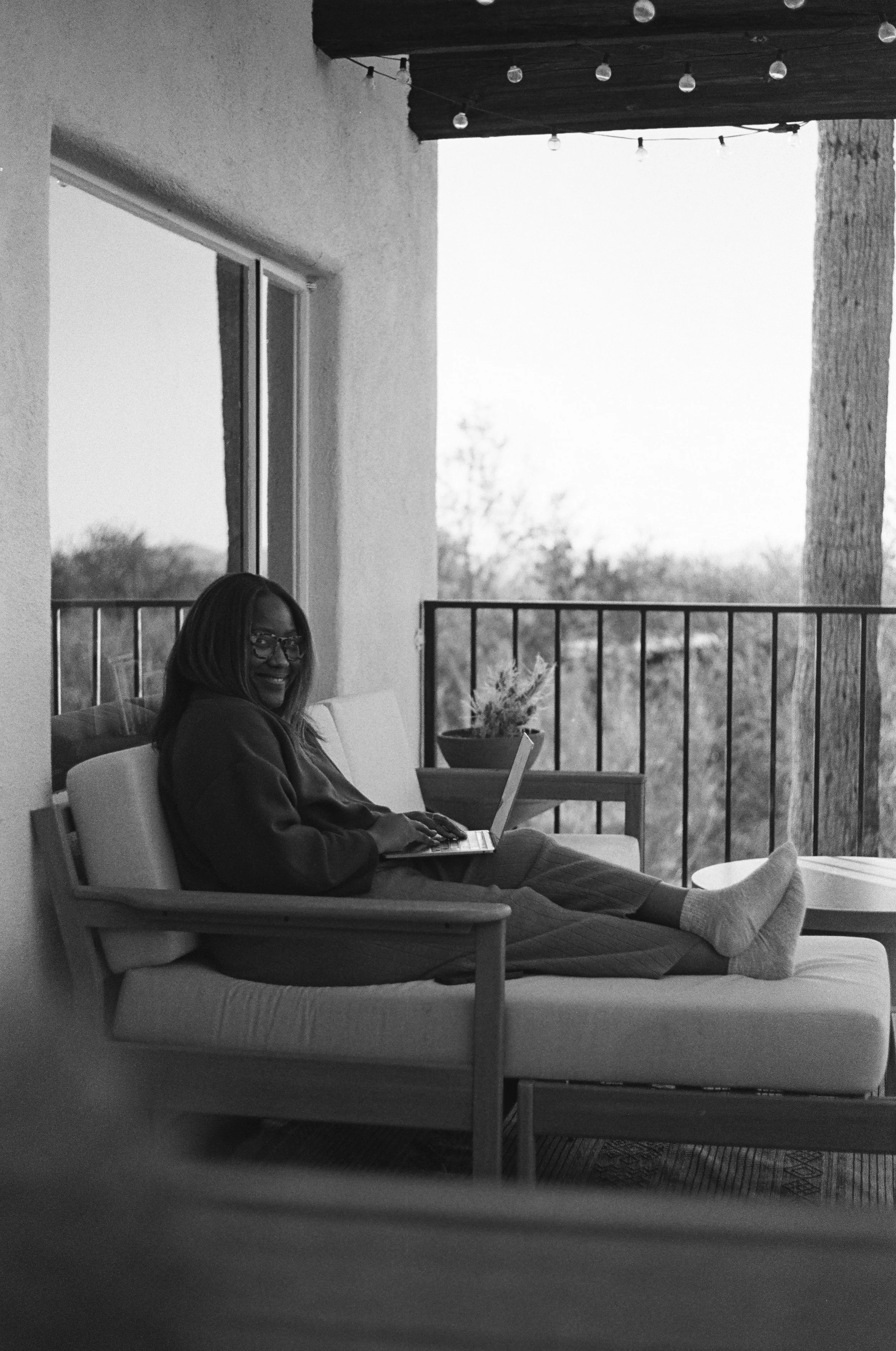 A woman sitting on a couch in a room, using a laptop. She is smiling and wearing glasses and a hoodie. The room has a large window, outdoor trees, a potted plant, and string lights overhead.