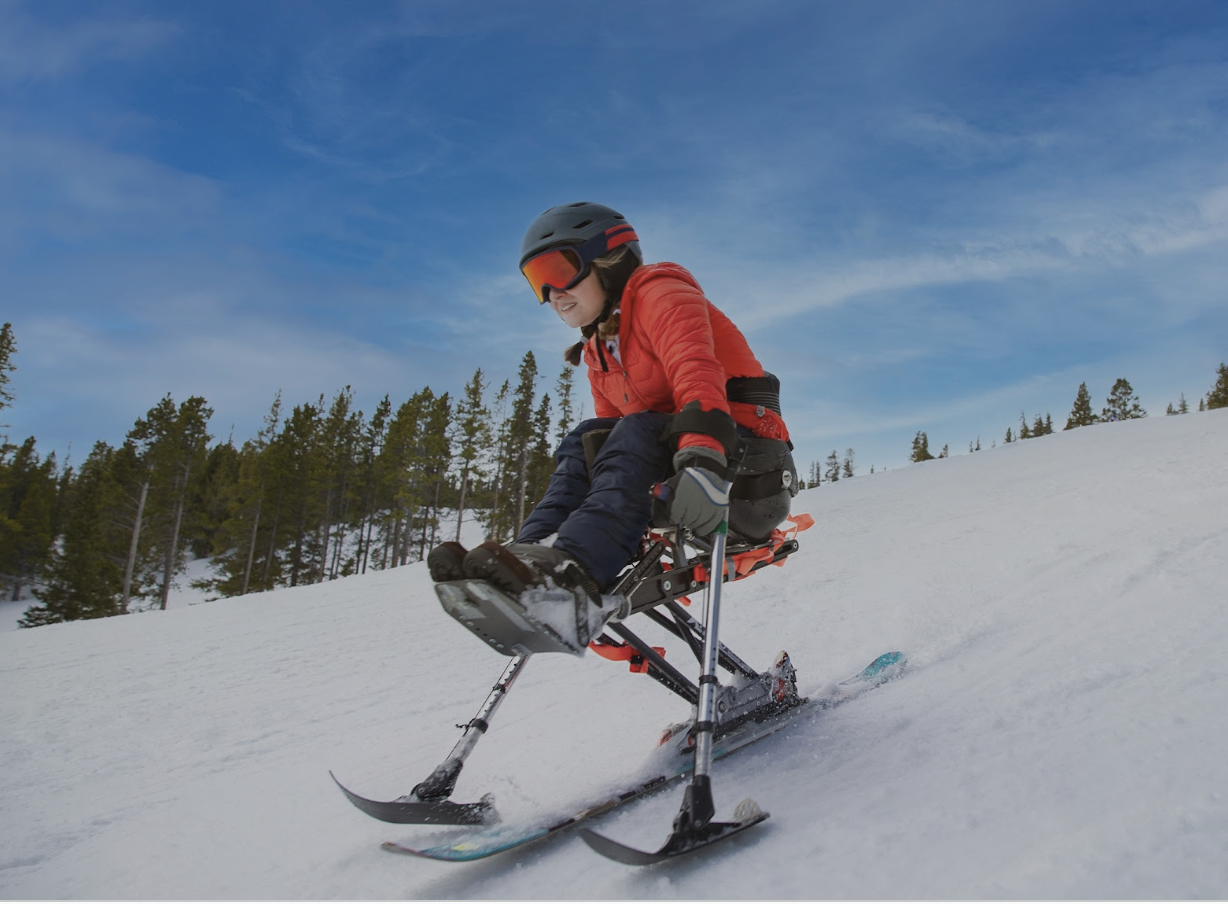 A person in a red jacket, black helmet, and ski goggles sitting on a sit-ski carving down a snowy slope with trees and blue sky in the background.