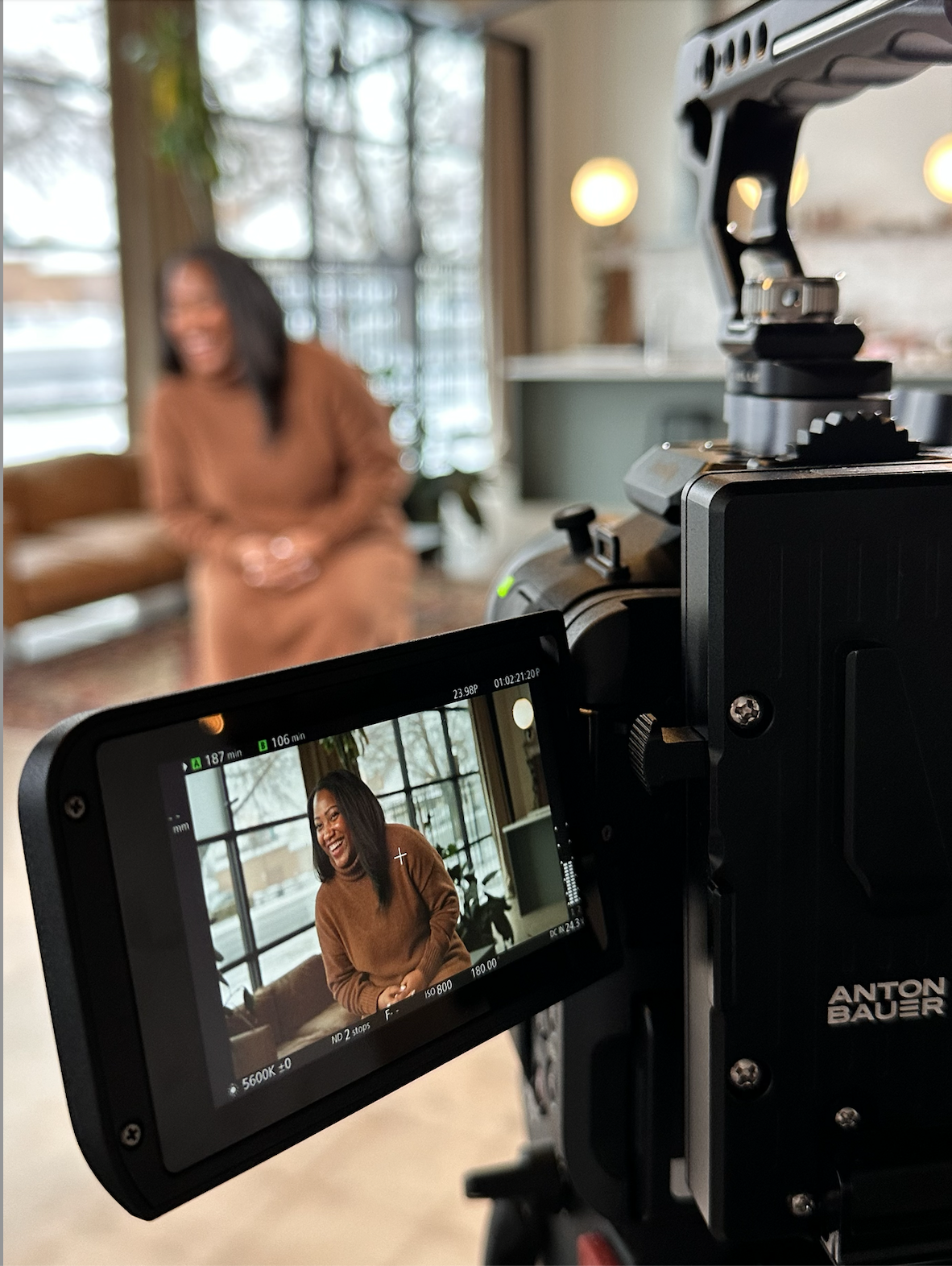 A camera recording a woman with shoulder-length dark hair, smiling and sitting on a sofa in a room with large windows, indoors.