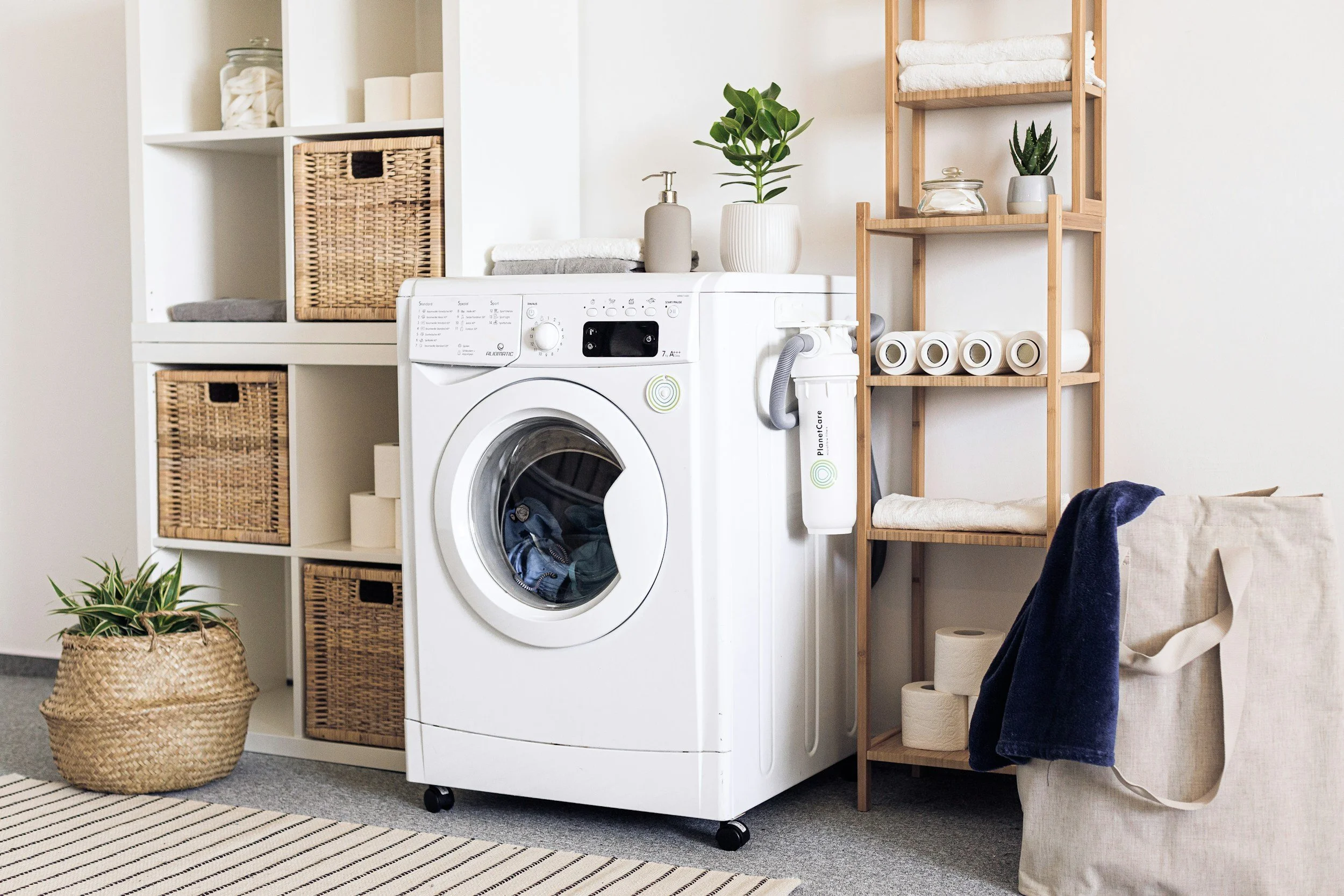 A laundry room with a white washing machine, wicker baskets, a potted plant, folded towels, and rolls of paper towels on a wooden shelf.