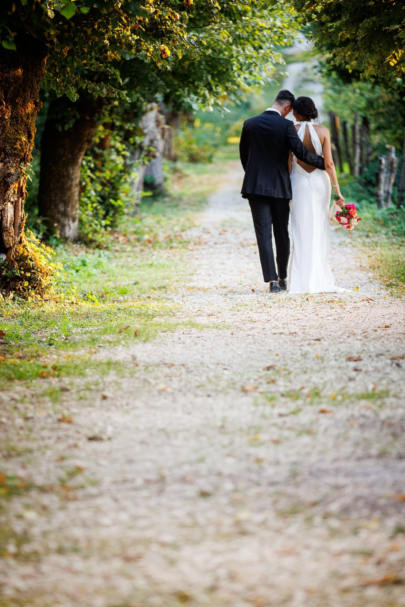 Un couple en mariée et marié marche dans un chemin de campagne bordé d'arbres verts. Photo : Samuel Coulon