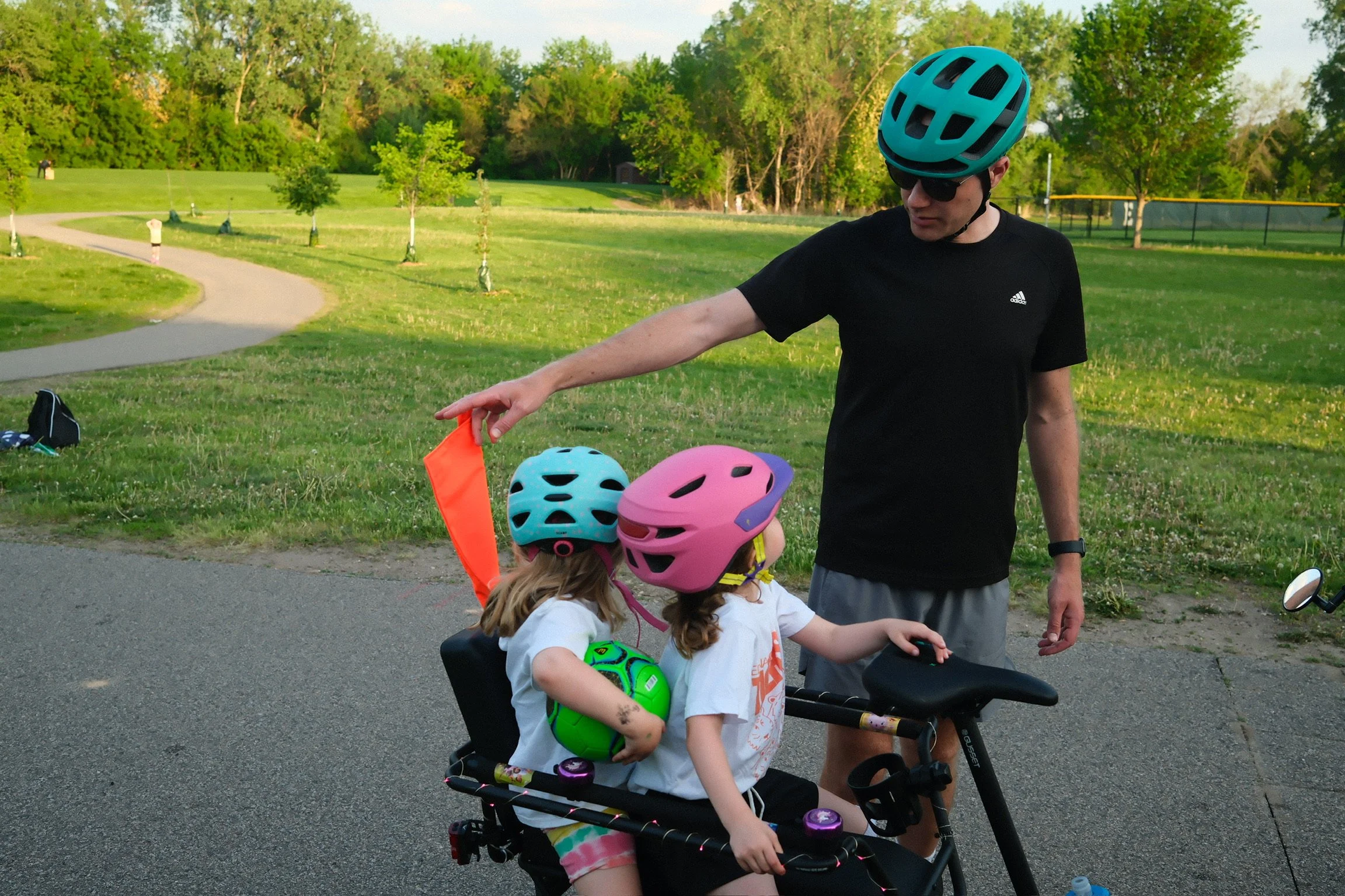A man wearing a blue helmet and sunglasses is giving a safety instruction to two young girls sitting on a bike, with both girls wearing pink helmets, in a park with green trees and a walking path in the background.