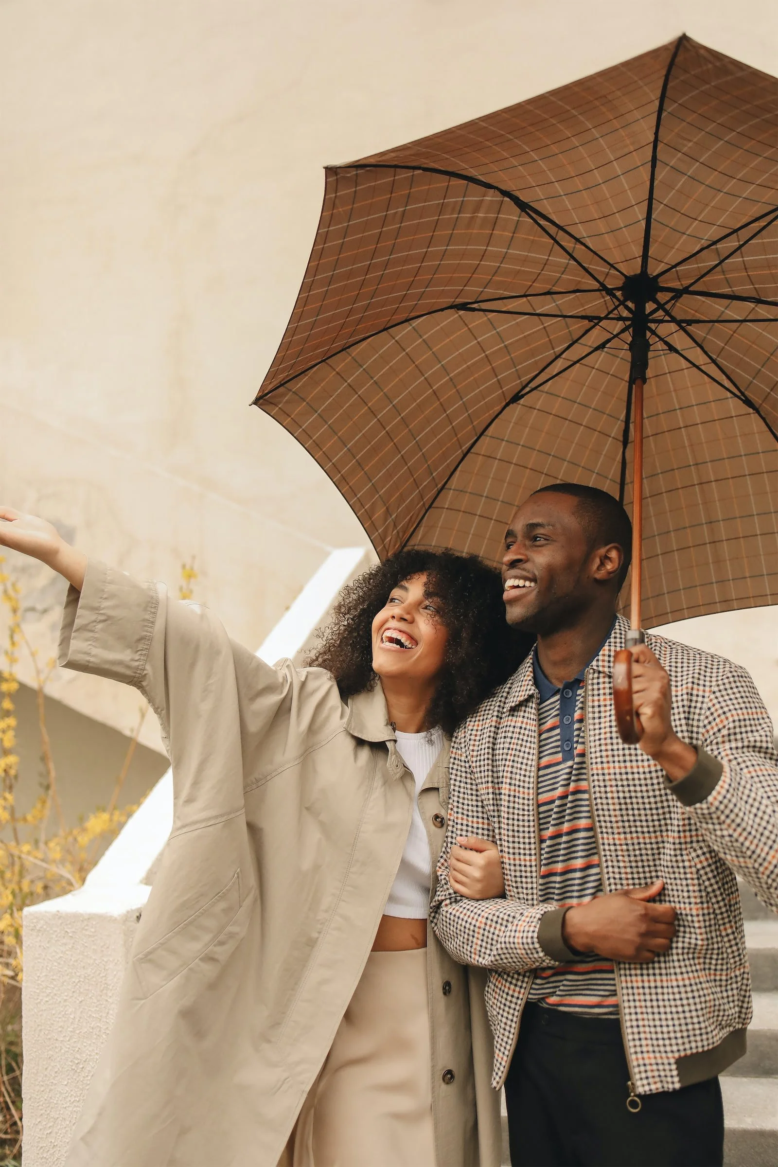 A smiling couple standing outside holding an umbrella and pointing at something, enjoying a rainy day.