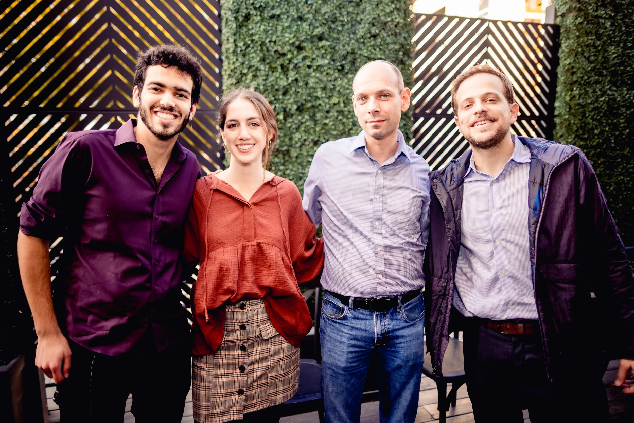 Four people smiling and posing outdoors in front of a decorative green wall and lattice privacy screen. From left to right: man with dark curly hair in a purple shirt, woman with light brown hair in a red blouse and checked skirt, bald man in a light blue checked shirt, man with short light brown hair in a purple jacket and checked shirt.