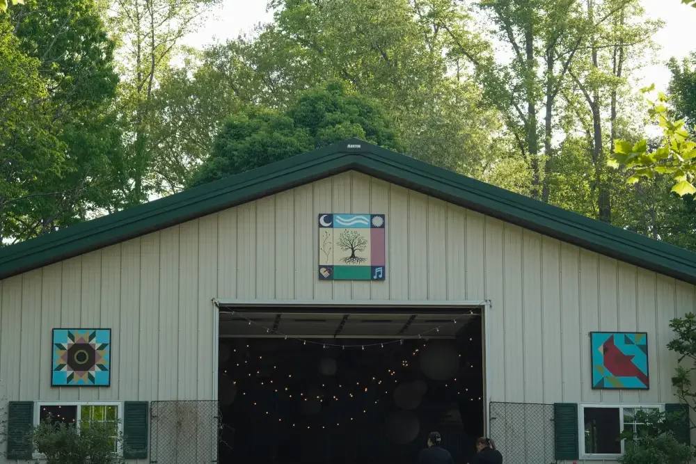 A barn with artwork on its exterior walls, two people are entering, trees surrounding the barn, and string lights are hanging inside.