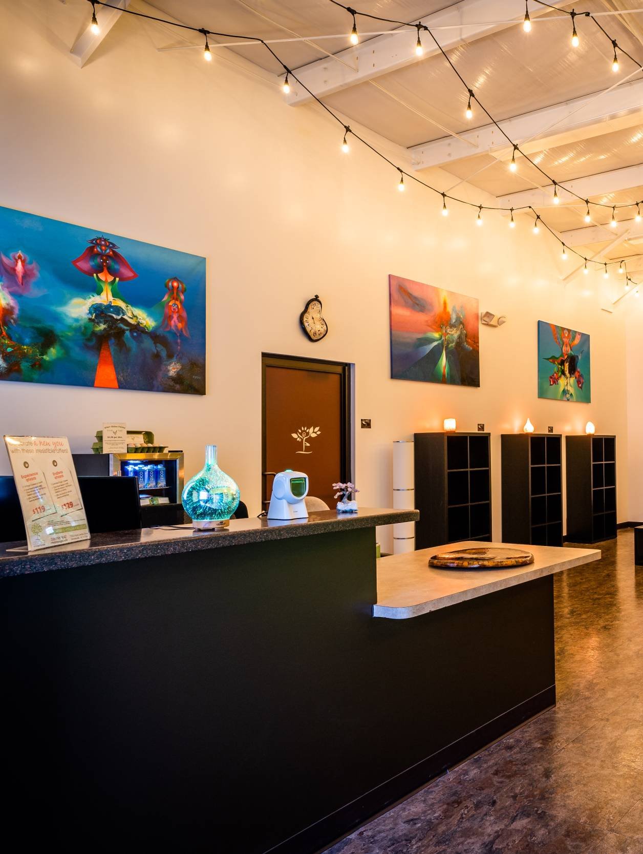 Gallery or reception area with paintings on the wall, warm lighting, a counter with decorative items and a digital device, black shelving units, and ceiling string lights.