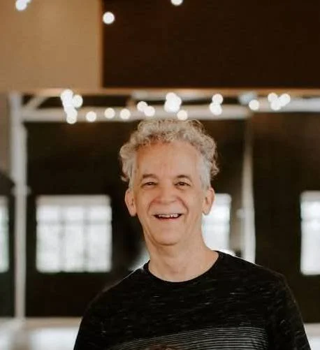 Smiling man with curly gray hair in a room with string lights hanging.