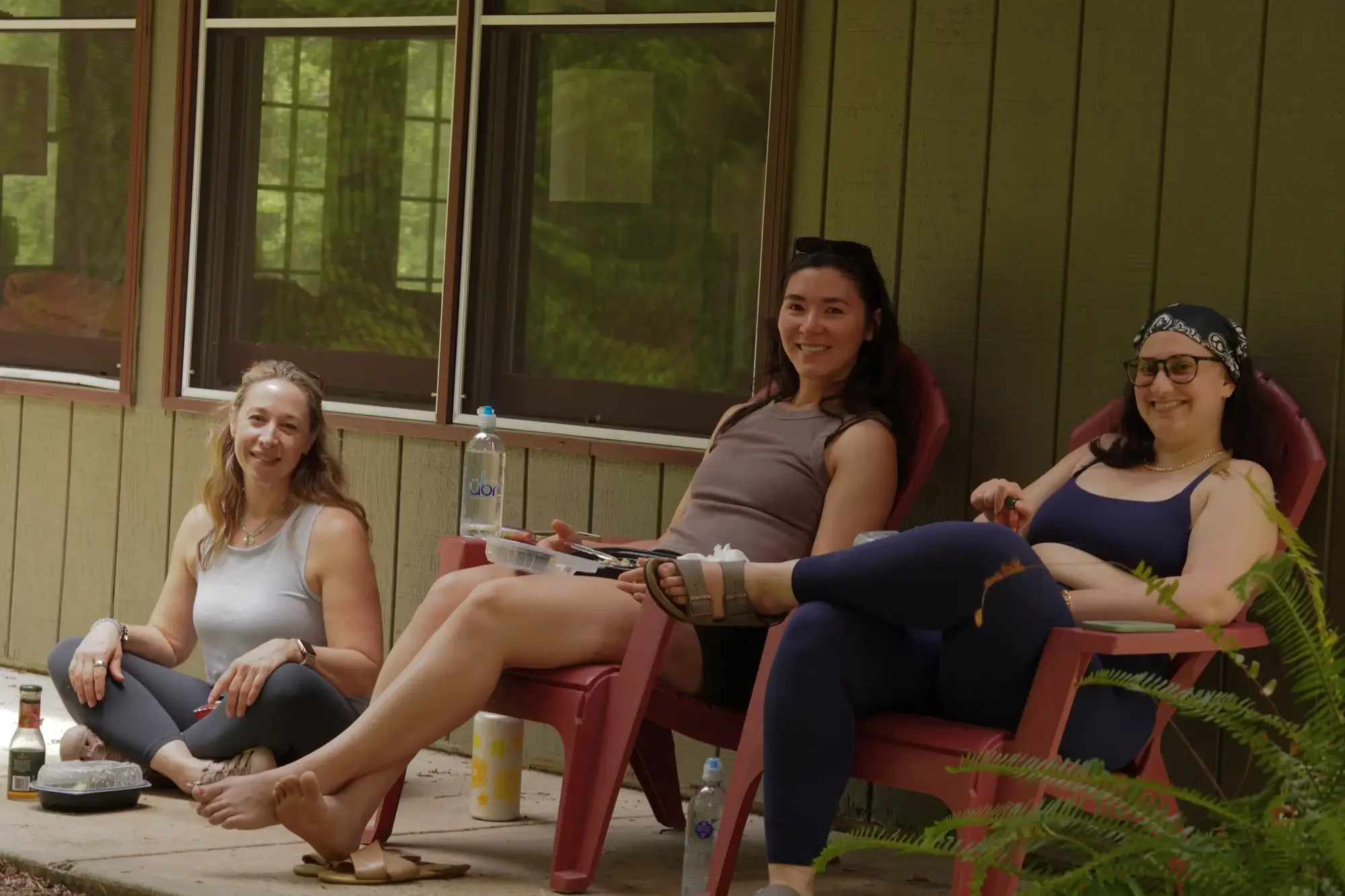 Four women sitting on patio chairs outside a house with green siding and windows, smiling and relaxing. One woman has long hair, wearing a white top; another has dark hair and sunglasses; another wears a bandana and glasses; the last woman has long c