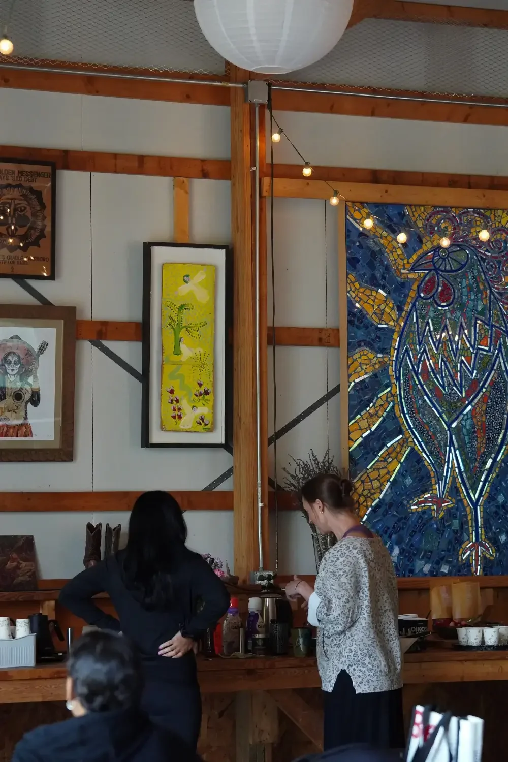 Two women in a café, one with dark hair and the other with light brown hair, standing at the counter. The wall behind has various colorful framed artworks, including a large mosaic of a bird.
