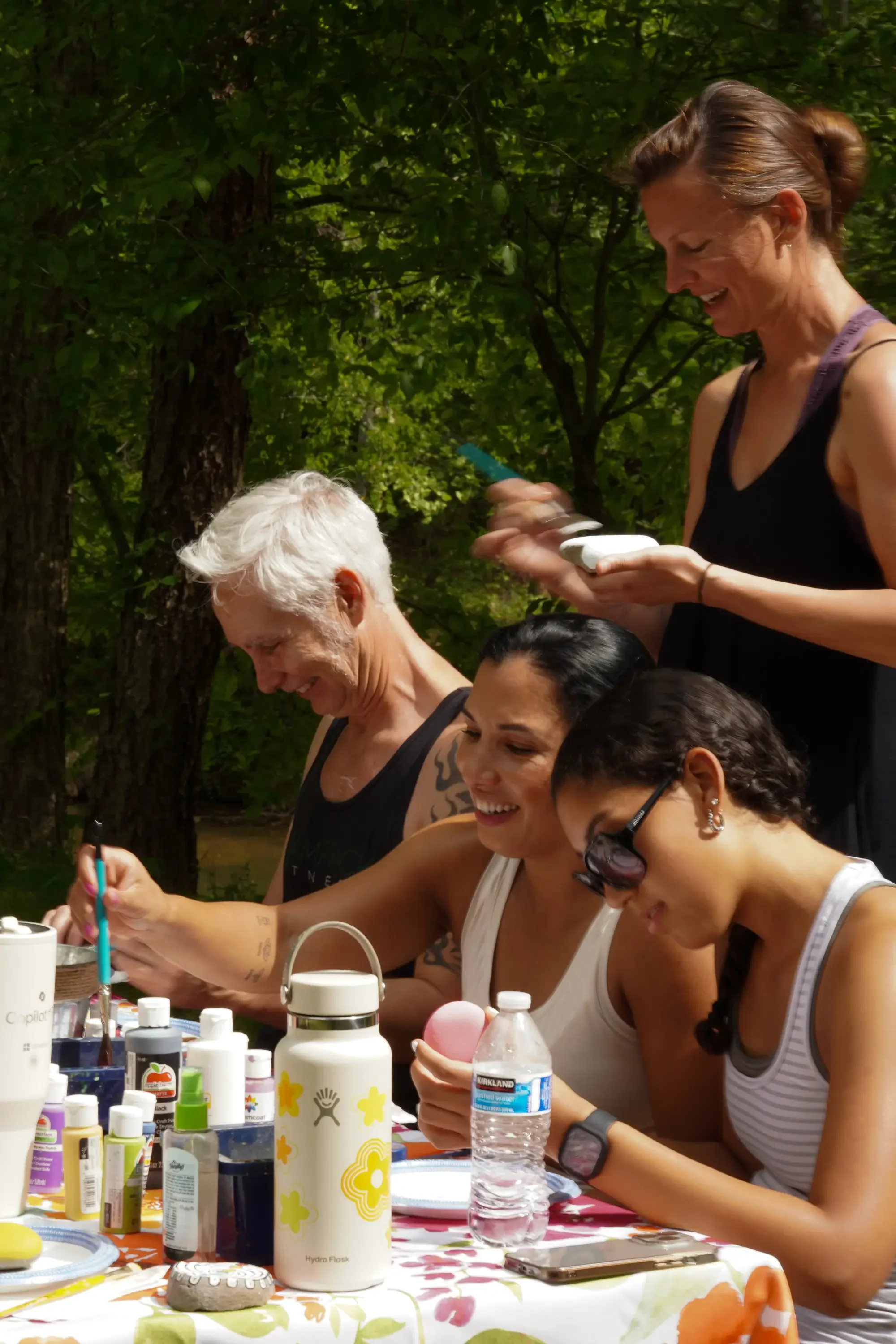 A group of women seated at an outdoor table with various art supplies, smiling and engaging in a creative activity, surrounded by trees and greenery.
